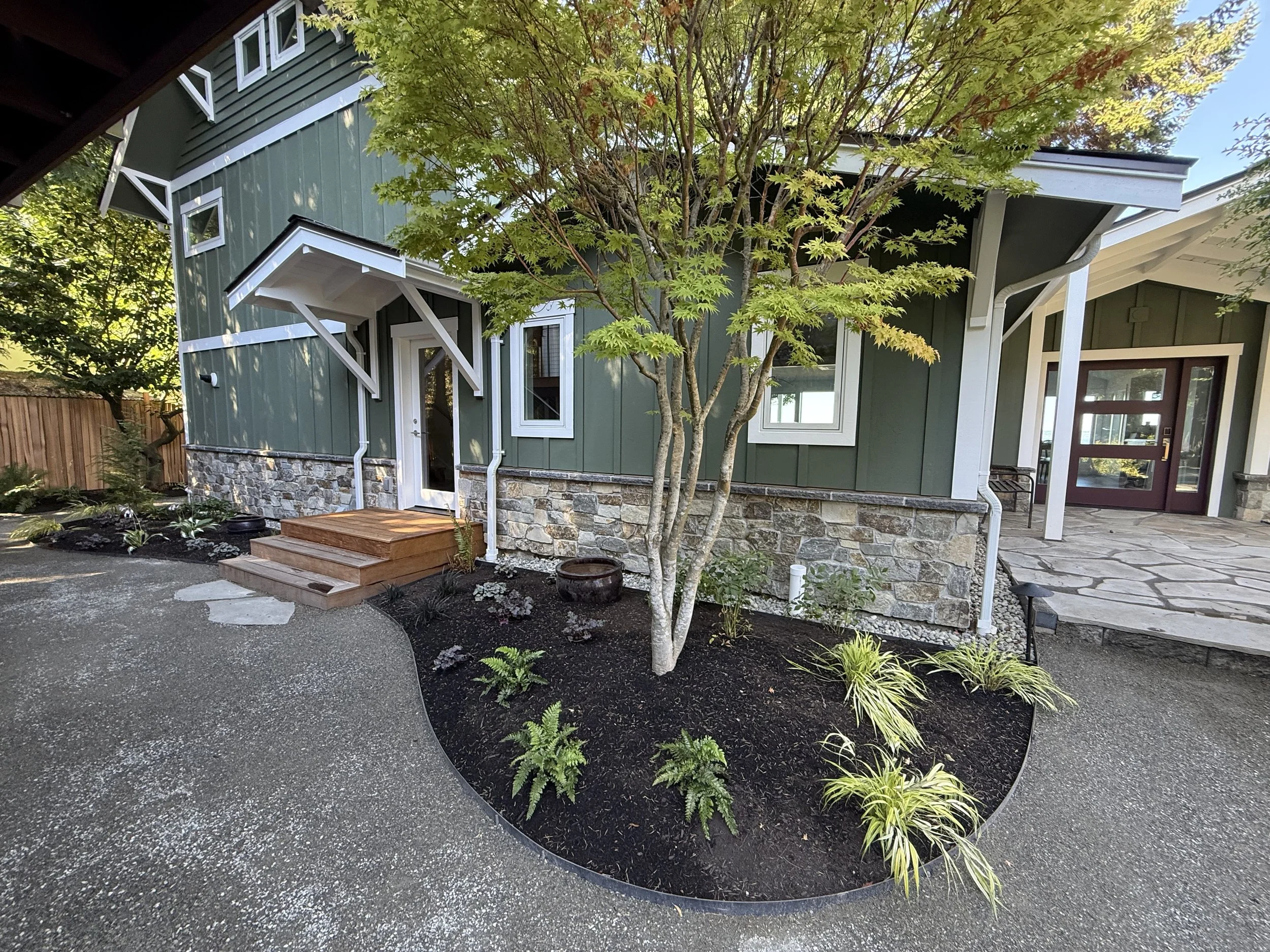 A modern house with green vertical siding, white trim, and stone foundation, surrounded by a landscaped yard with plants, trees, and a stone path.