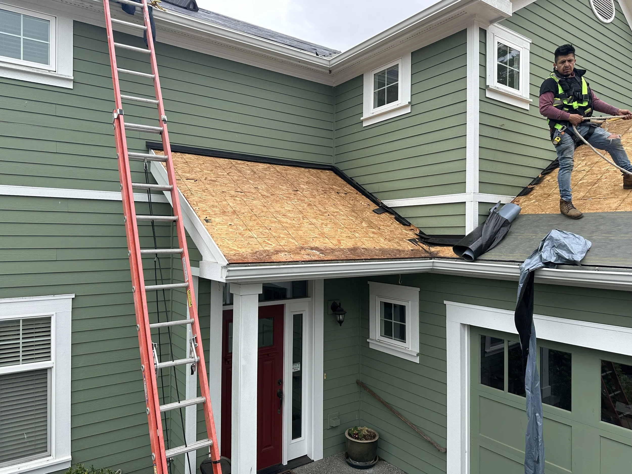 A man in work clothes standing on a roof that is under construction, with orange siding, a red door, and green exterior walls of a house, next to a ladder and construction materials.