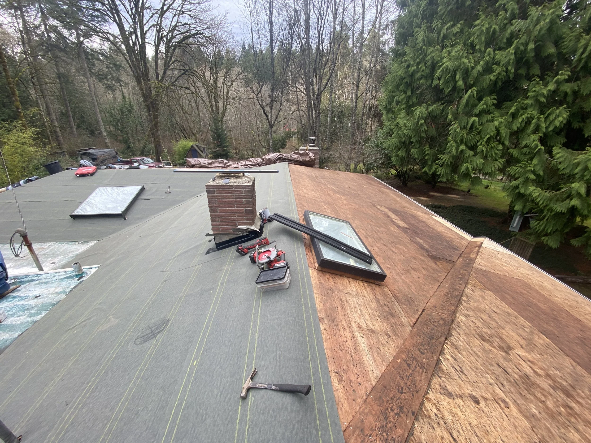 Roof of a house under construction or renovation, with tools and materials scattered, and trees in the background.