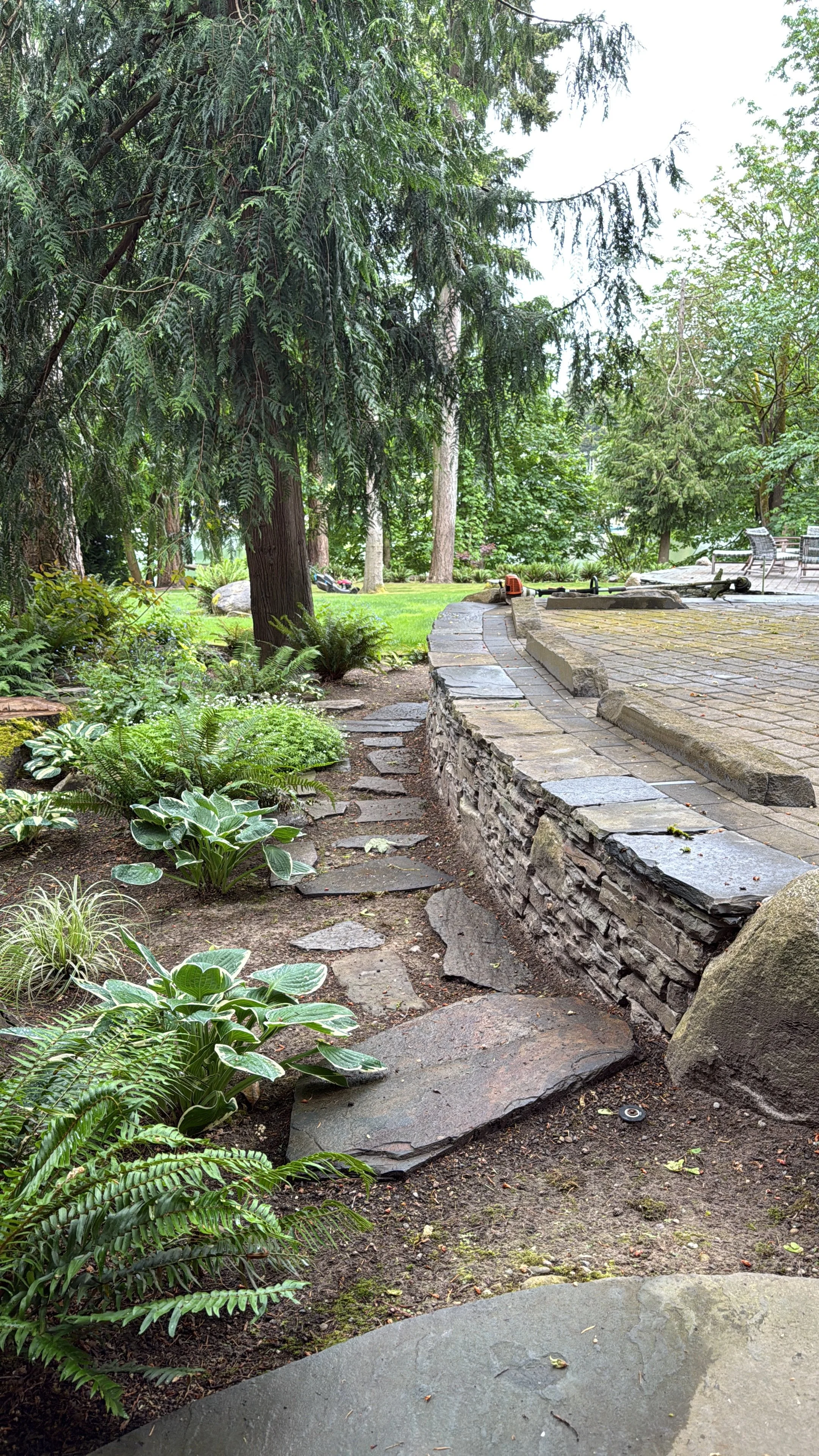 A landscaped garden with tall trees, green shrubs, a stone pathway, and outdoor furniture in the background.
