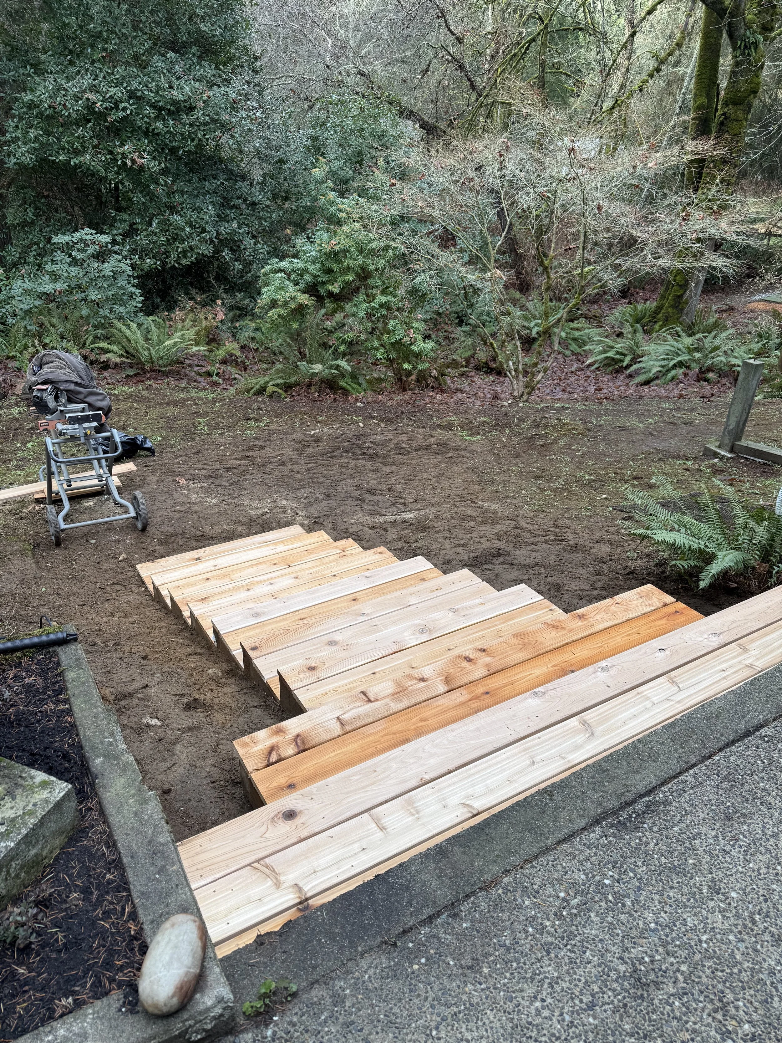 The image shows a wooden walkway under construction in a forested area, with construction tools and a backpack on the ground nearby, surrounded by green plants and trees.
