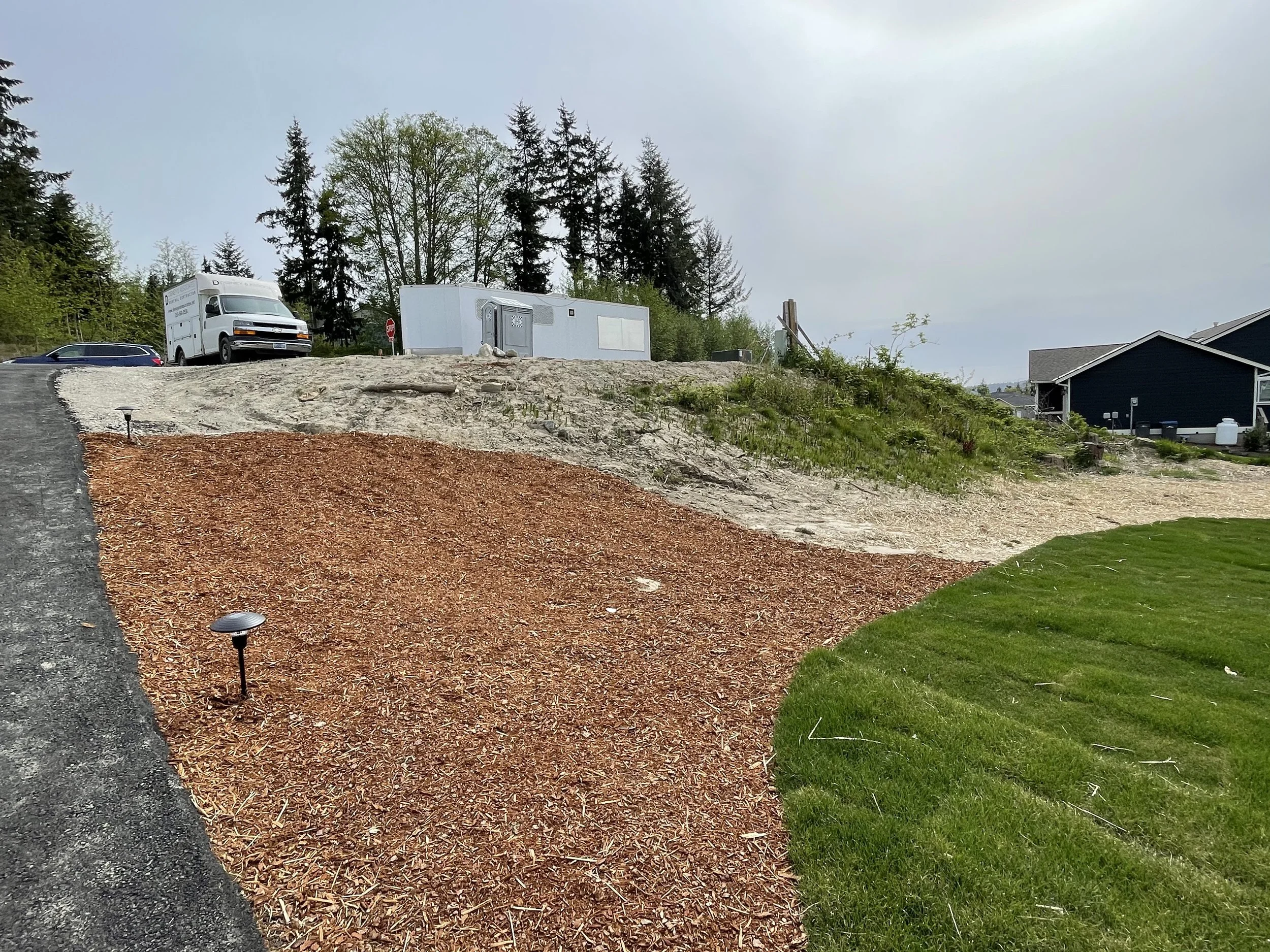 Yard with a curved pathway, mulch, grass, a row of solar lights, a parking lot with a white van, trees, a small blue building, and a house with a porch.