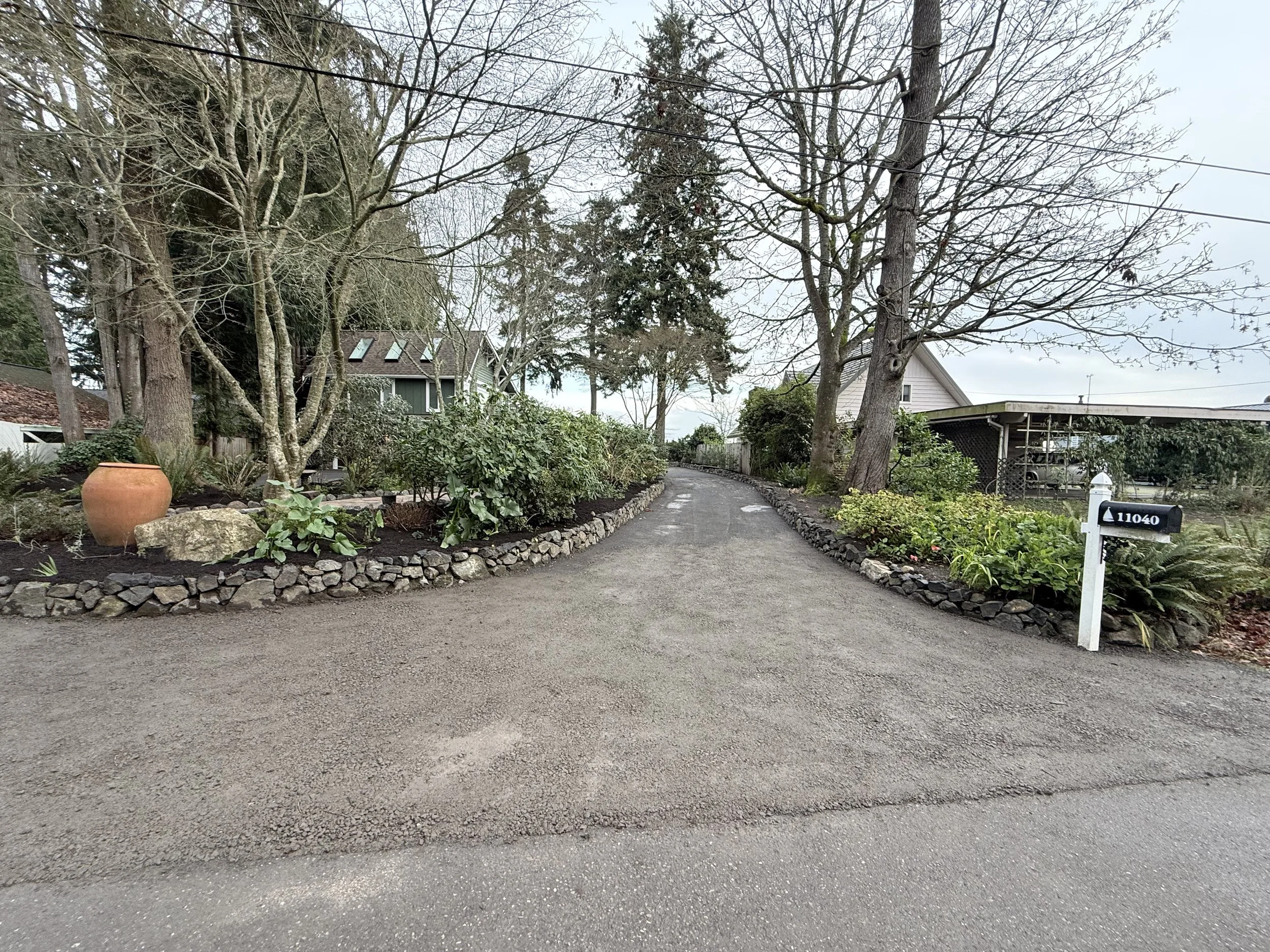 A gravel driveway curving through a garden with trees, shrubs, a large ceramic pot, and a white mailbox with the number 11040, under a cloudy sky.
