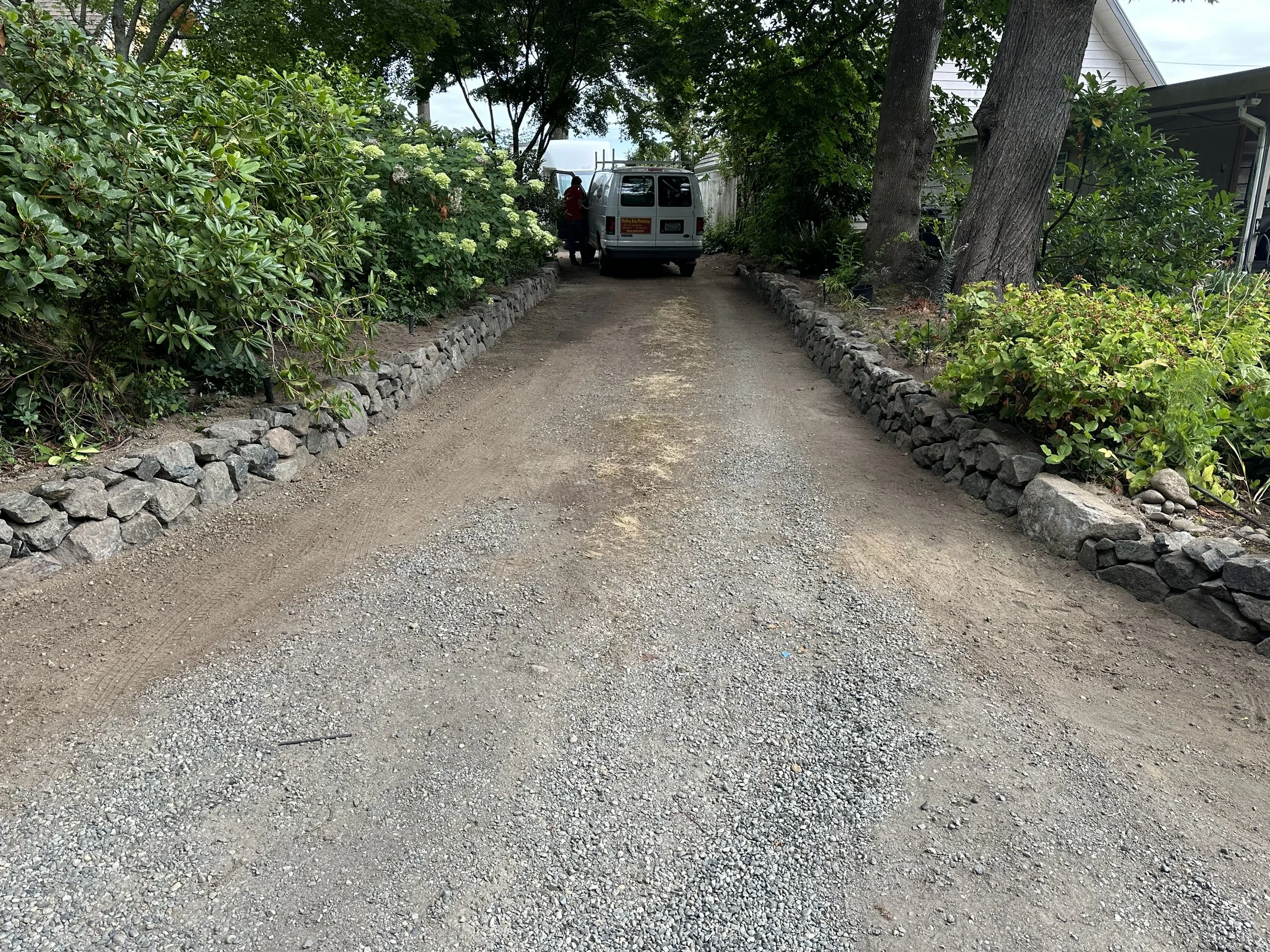 A dirt driveway with stone borders on both sides, surrounded by green bushes and trees, with a parked white van and a person nearby in a residential area.