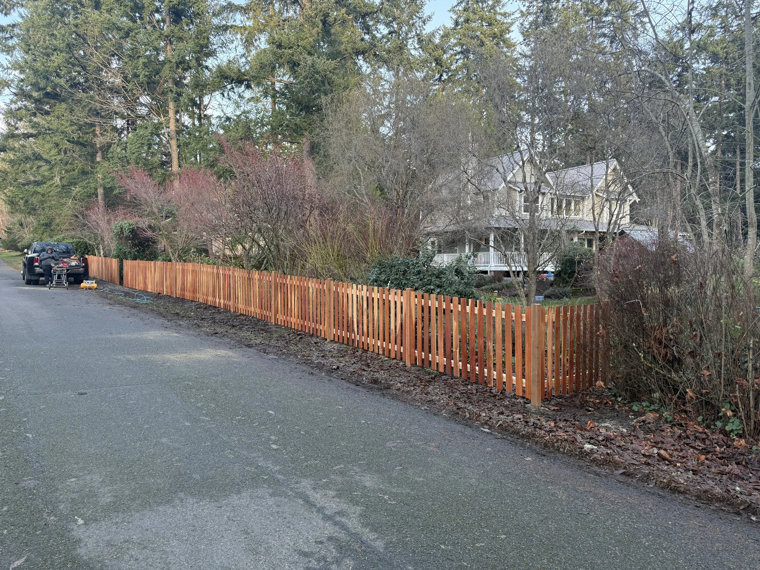 A residential street with a new wooden fence, a black pickup truck parked on the side, and a house surrounded by trees in the background, during daytime.