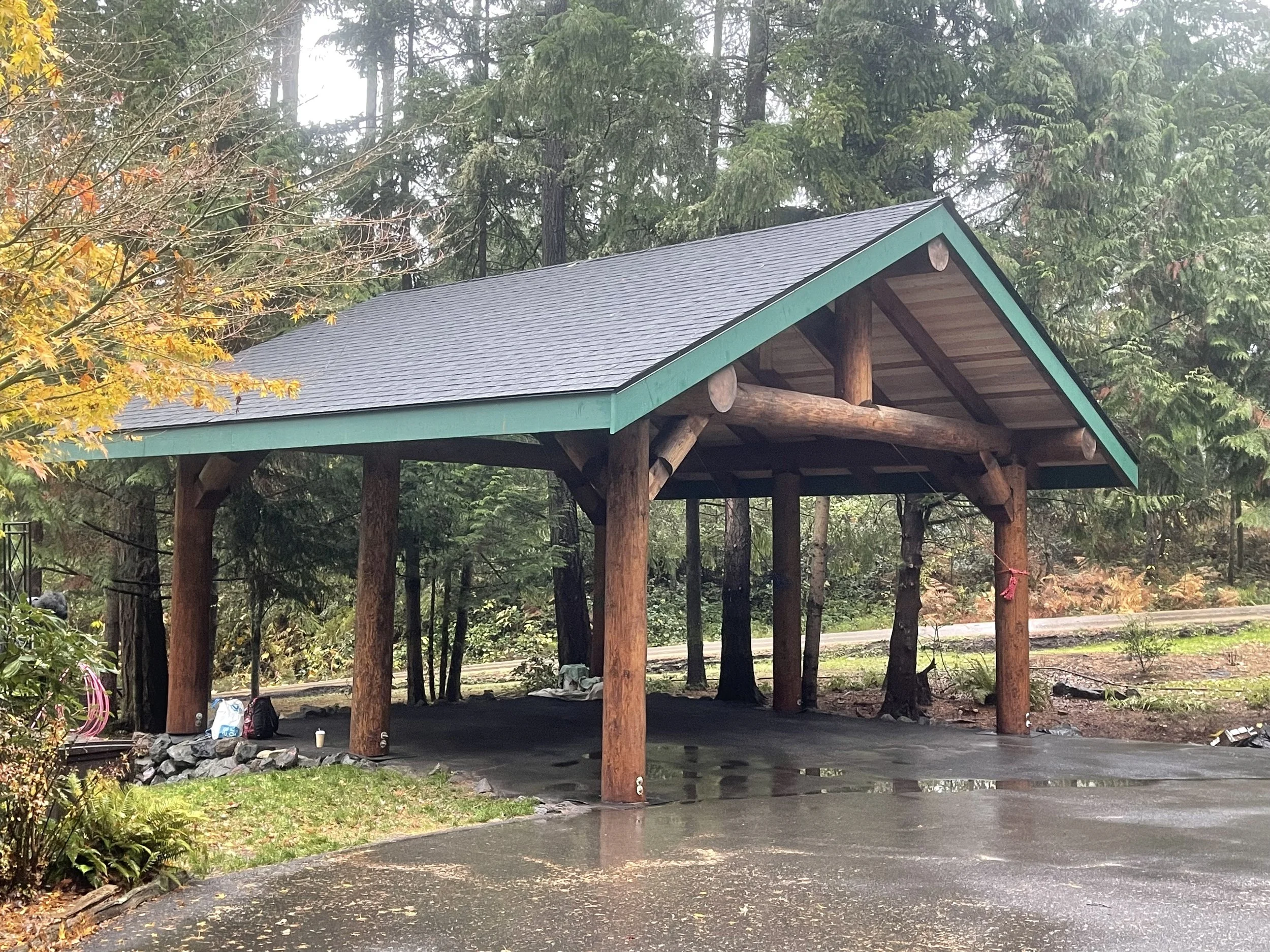 A wooden structure with a dark gray shingled roof, supported by large logs, situated in a forest. The background consists of tall green trees with some autumn-colored leaves, and there is a driveway or paved area in front of the structure.