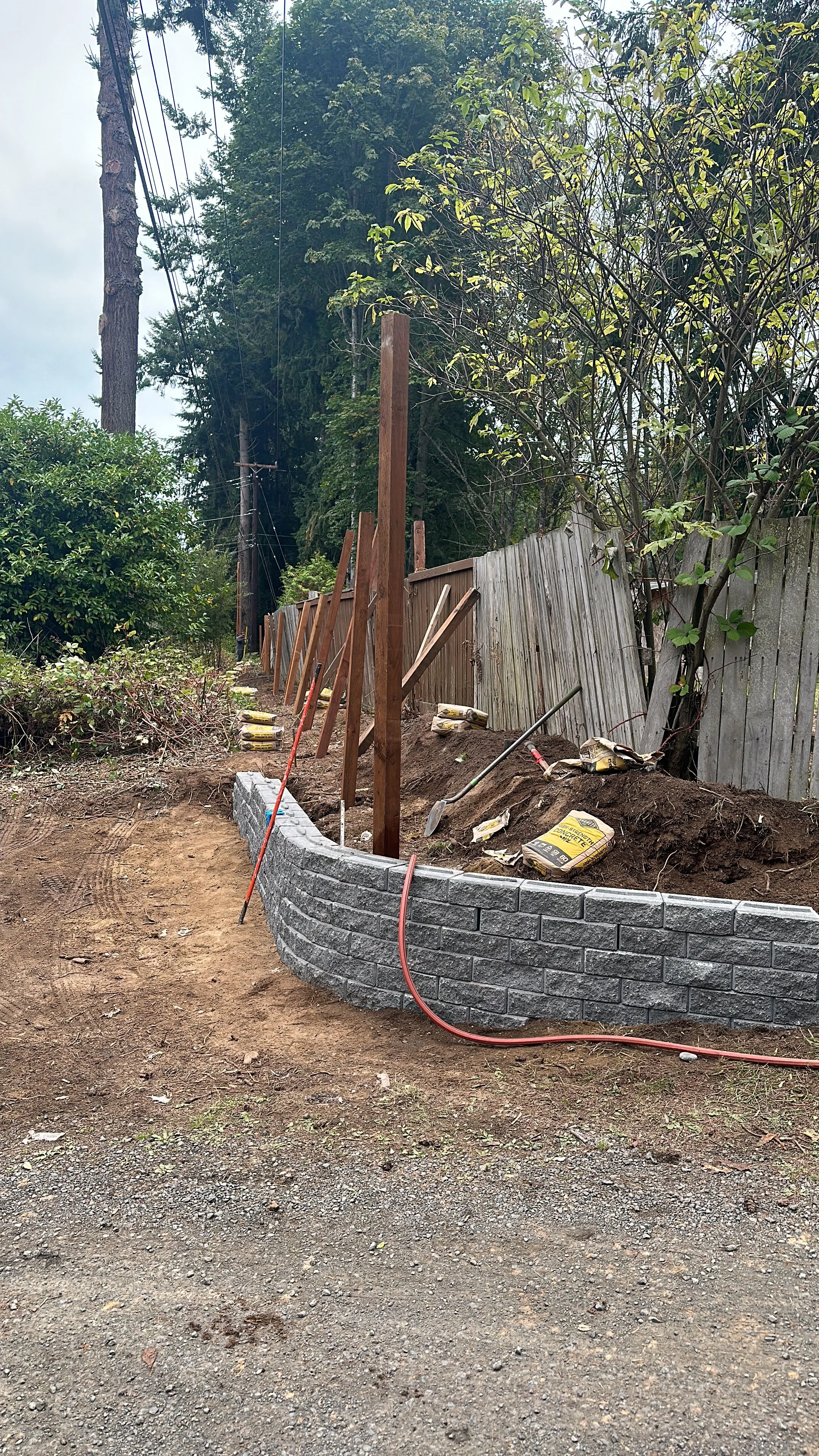 Construction site showing a newly built curved brick wall, with wooden supports, tools, and materials for landscaping or fencing in progress, surrounded by trees and a wooden fence.