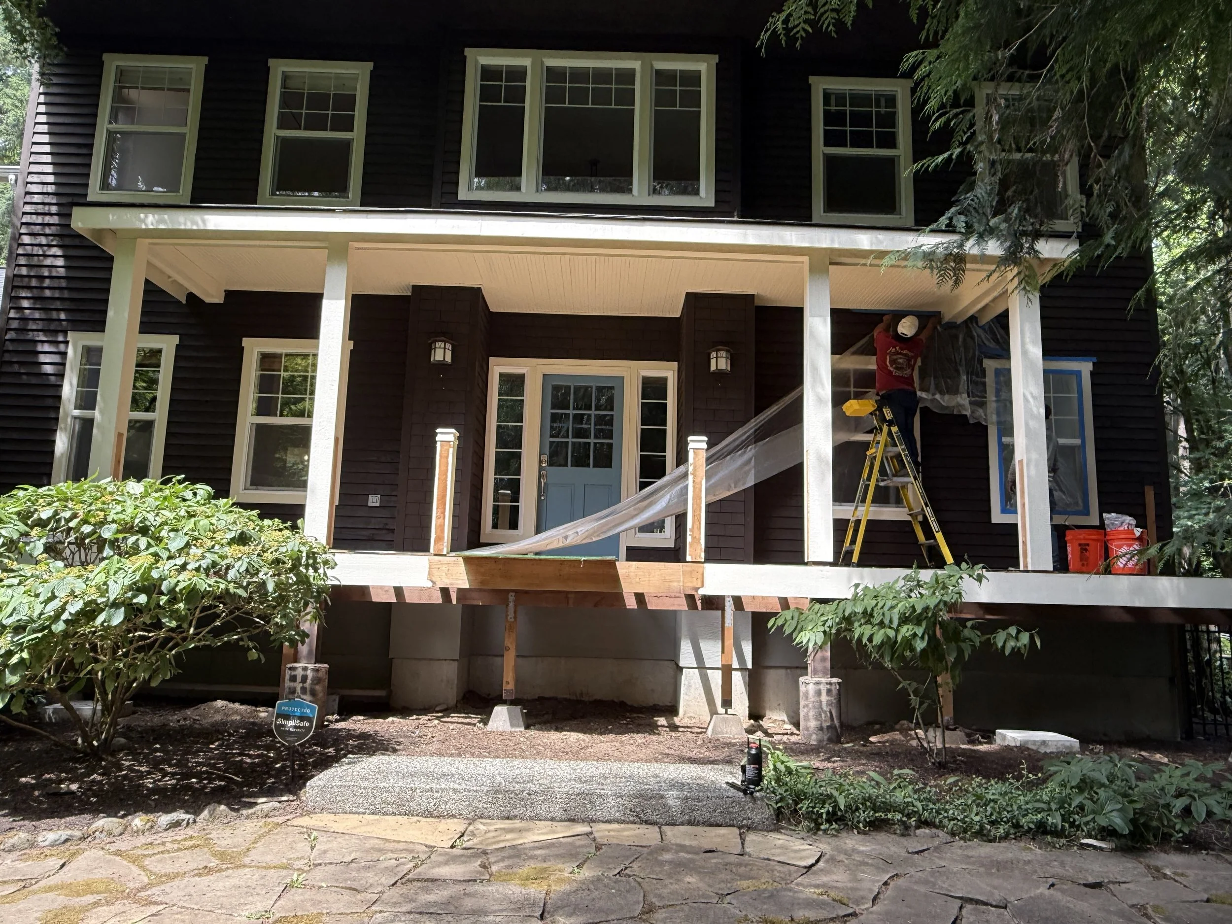 Front of a house under construction with an unfinished porch, a ladder, and a worker installing or repairing window trim, surrounded by greenery.