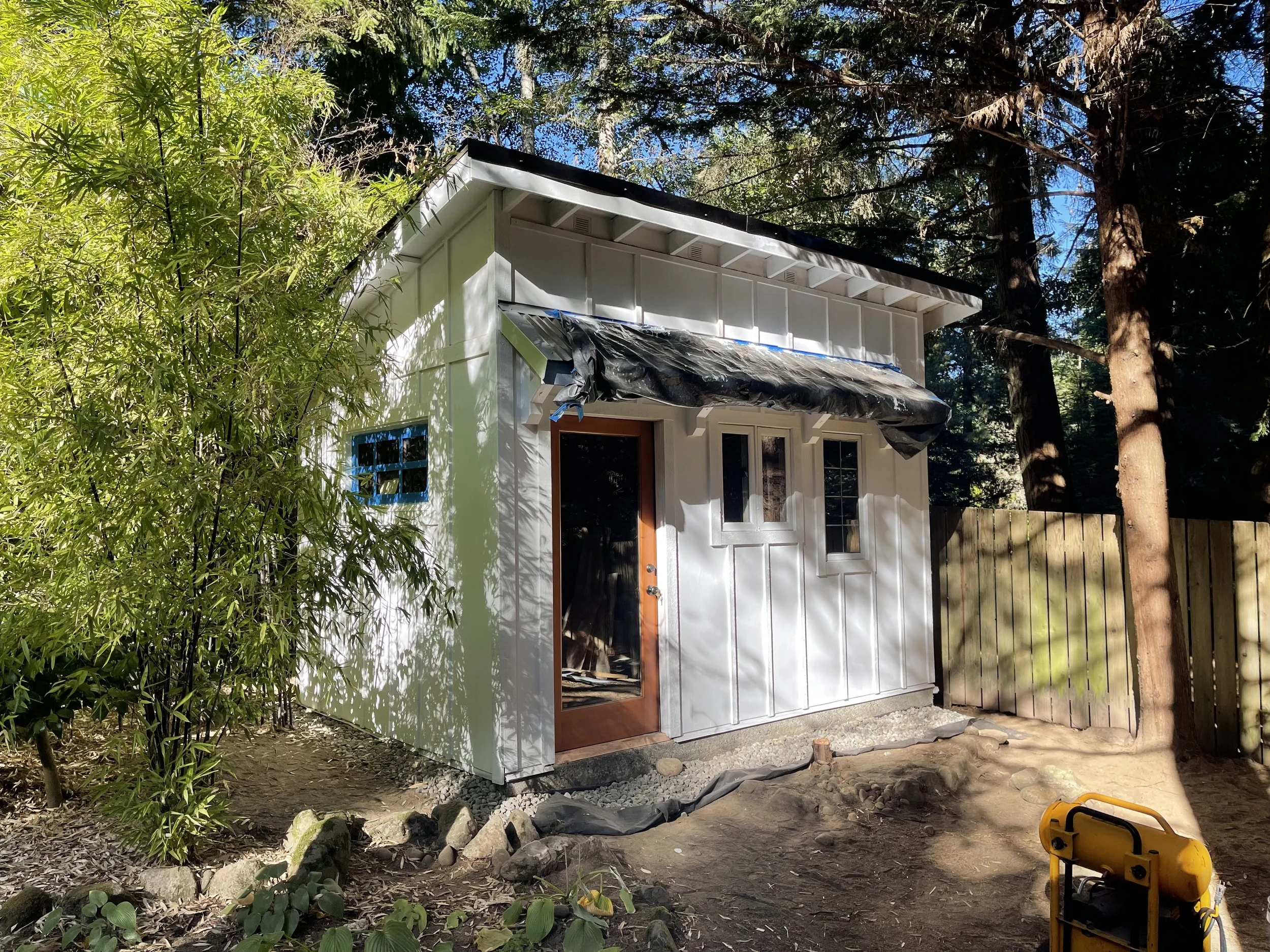 A small white house with black trim on the roof, surrounded by tall trees and green foliage. The house has a wooden door and window frames, and there appears to be construction or renovation work ongoing with materials and tools like a yellow power tool visible in the yard.