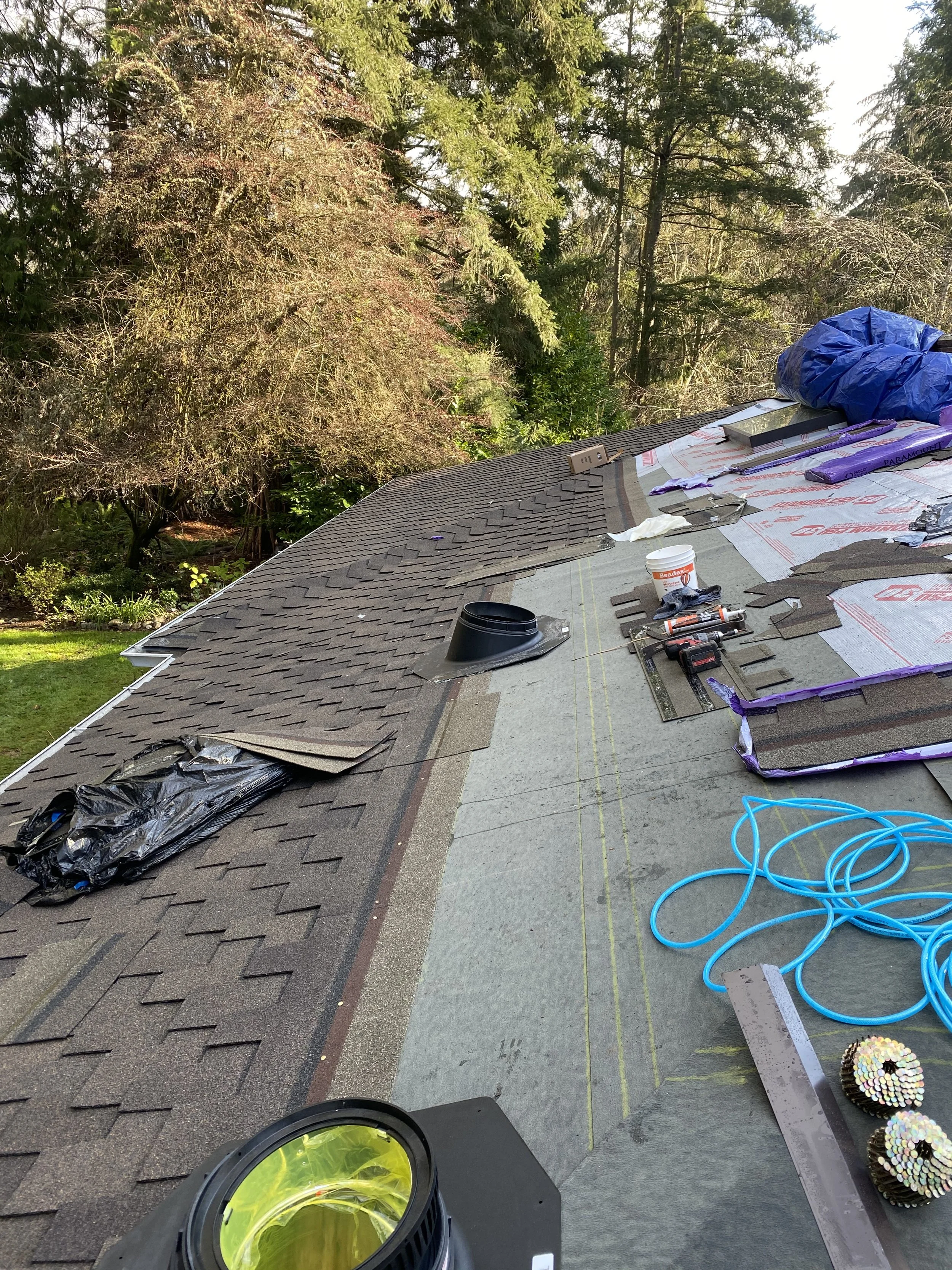 Photograph of a house roof under construction or repair, with roofing materials, tools, and equipment scattered on the surface, and surrounded by trees and greenery.