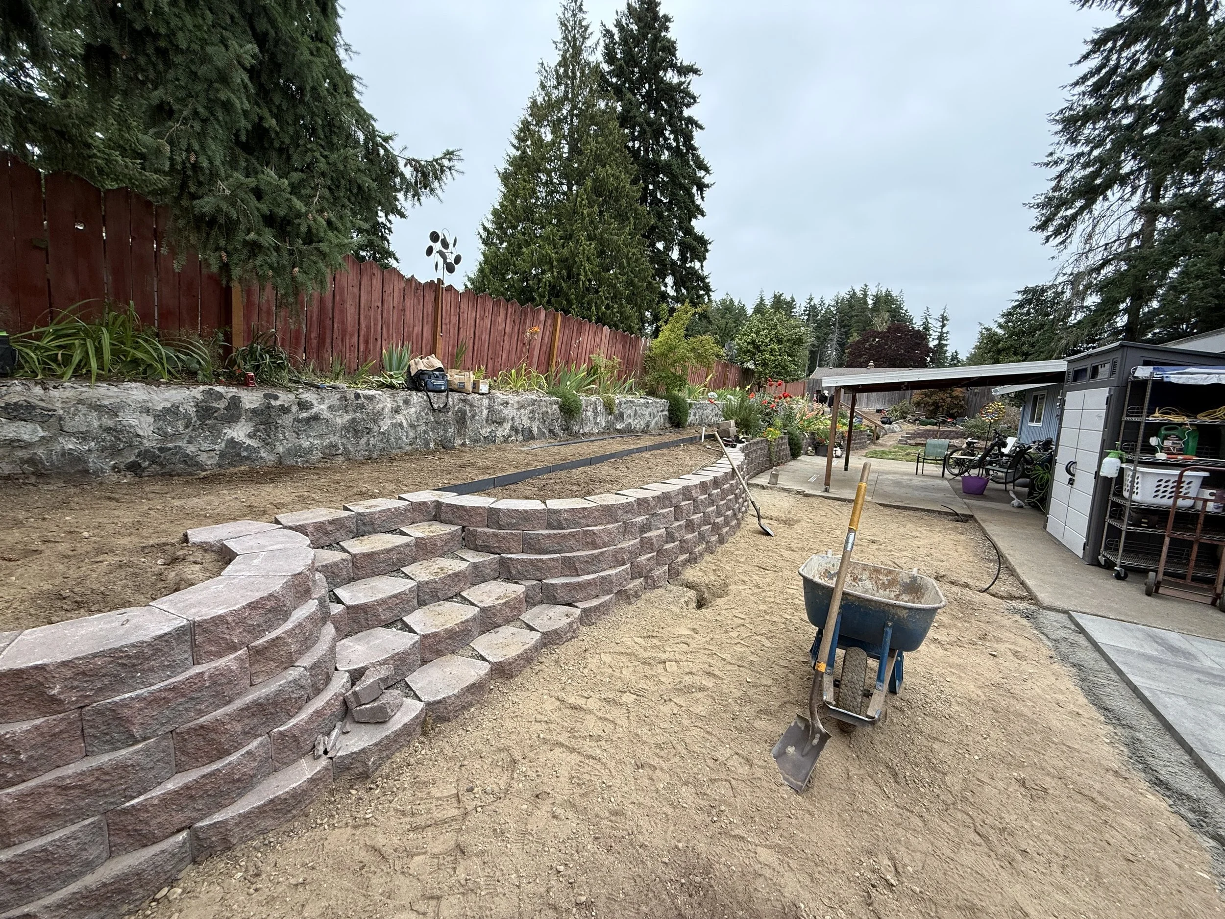 A backyard with a partially built stone retaining wall, a dirt area in the foreground, and various garden plants. There is a red wooden fence, tall trees, and gardening tools and supplies around the area.