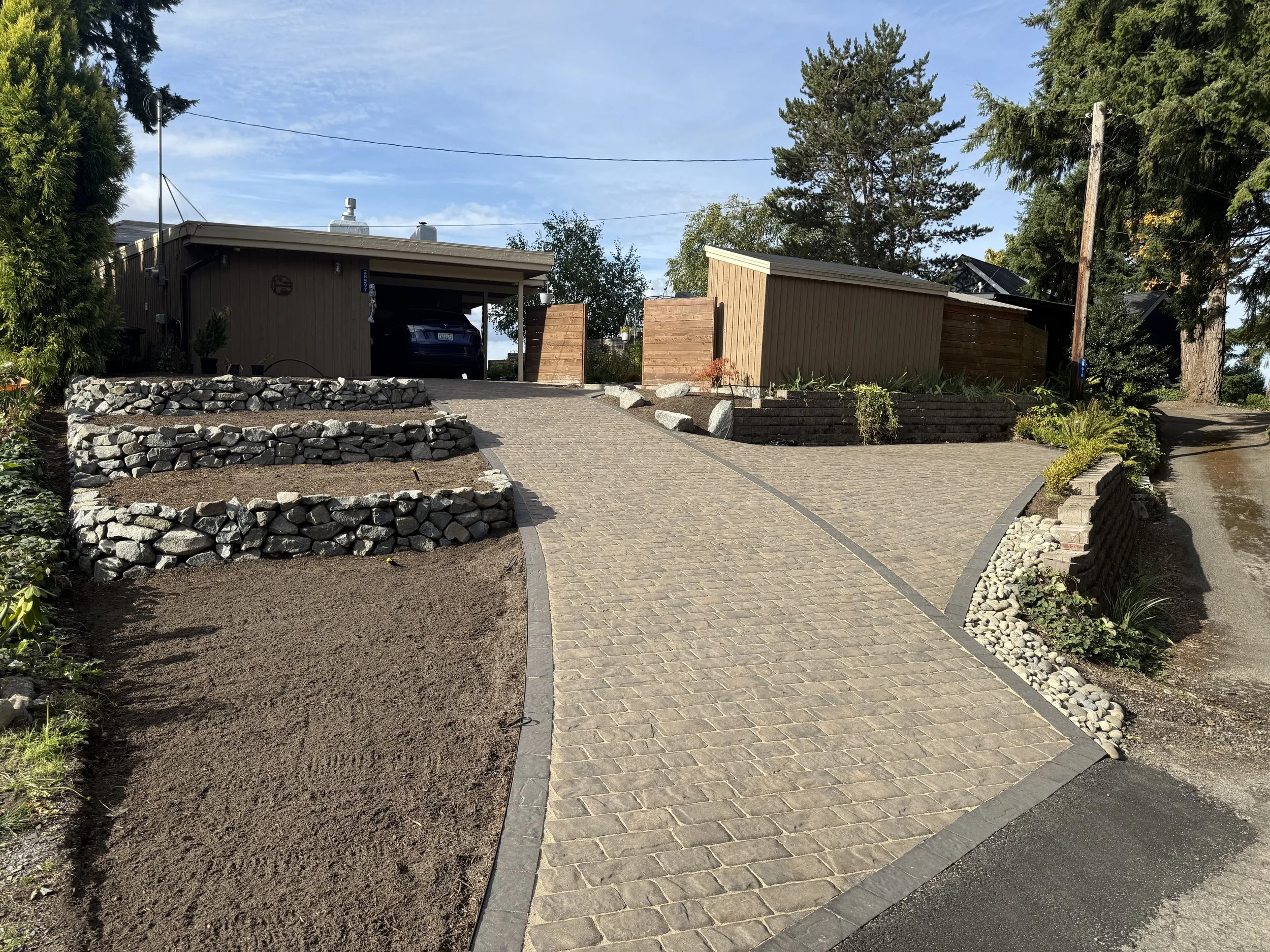 A landscaped outdoor area with a curved brick pathway, a new garden bed on the left with dark soil, and existing trees and houses in the background.