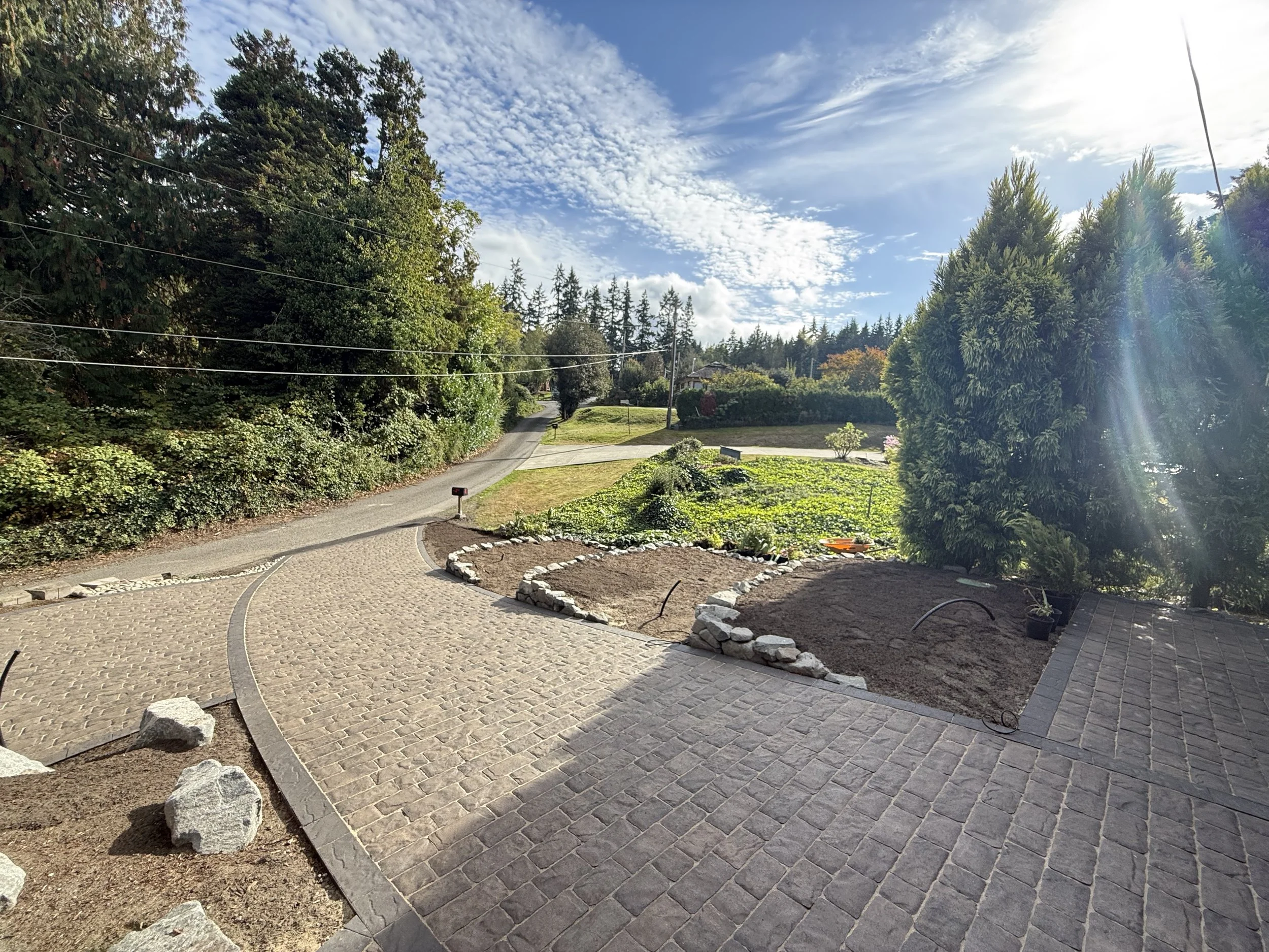 A residential neighborhood street with a driveway, green lawns, trees, and a sky with clouds and sunshine.