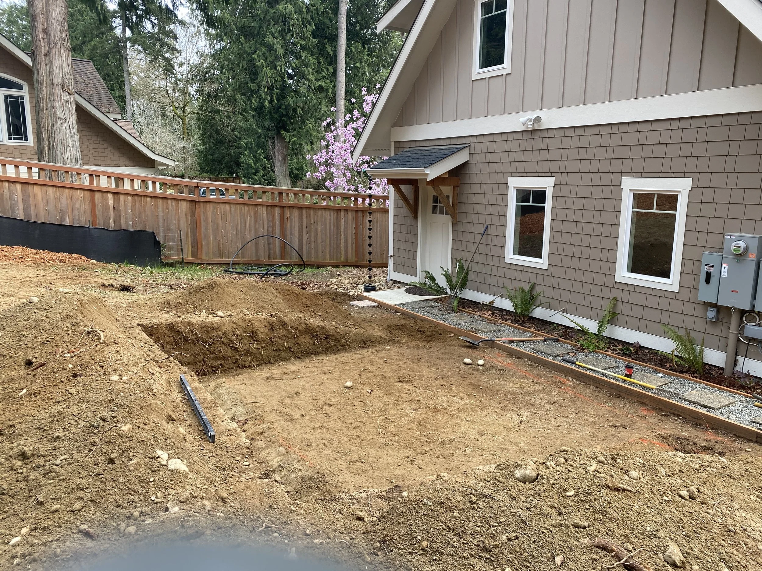 A backyard with dirt and construction materials in the foreground, a new fence, a house with beige siding, and trees in the background.