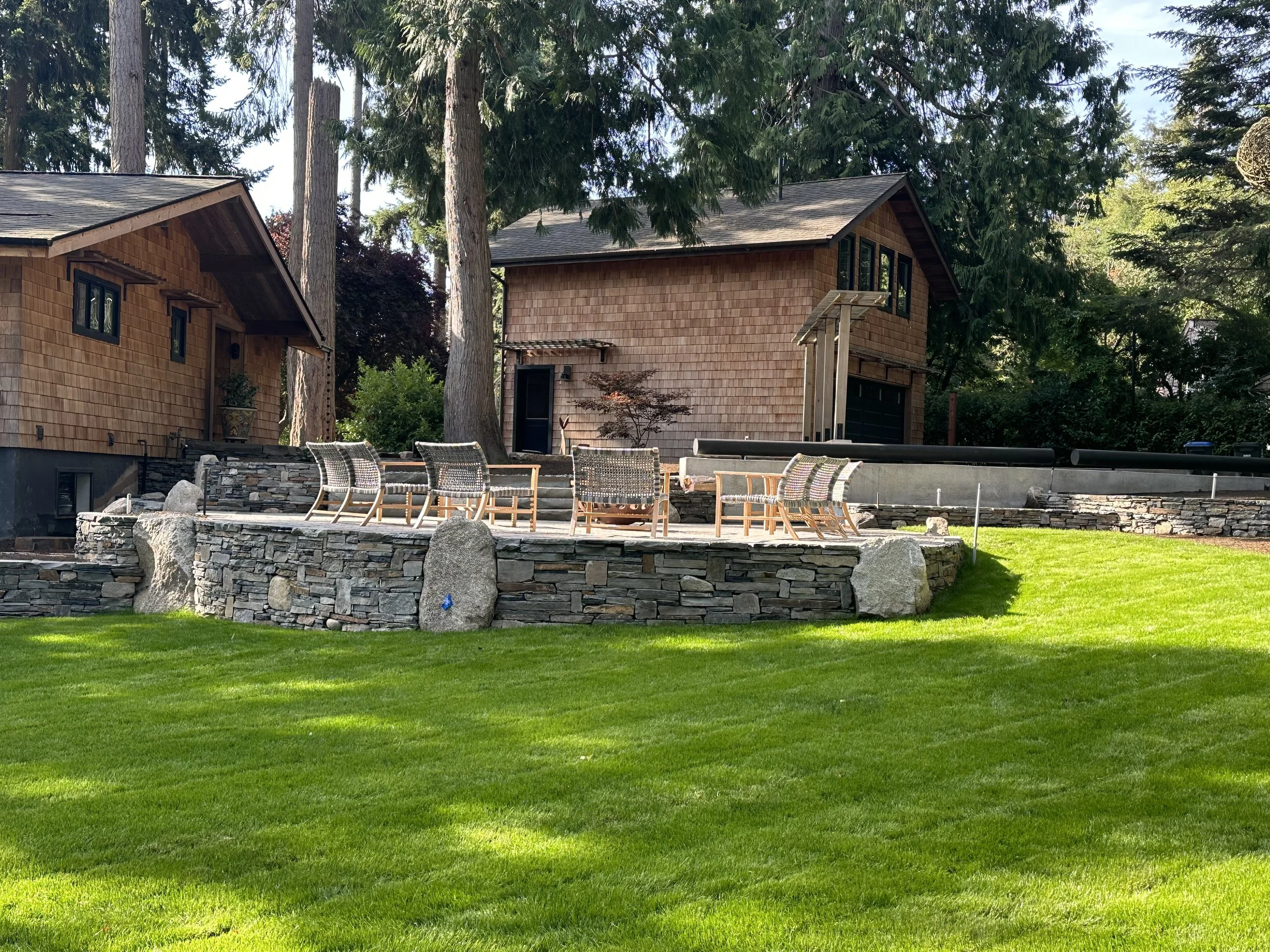 A backyard with a stone patio, organized outdoor furniture, mature trees, and a house with wooden siding.