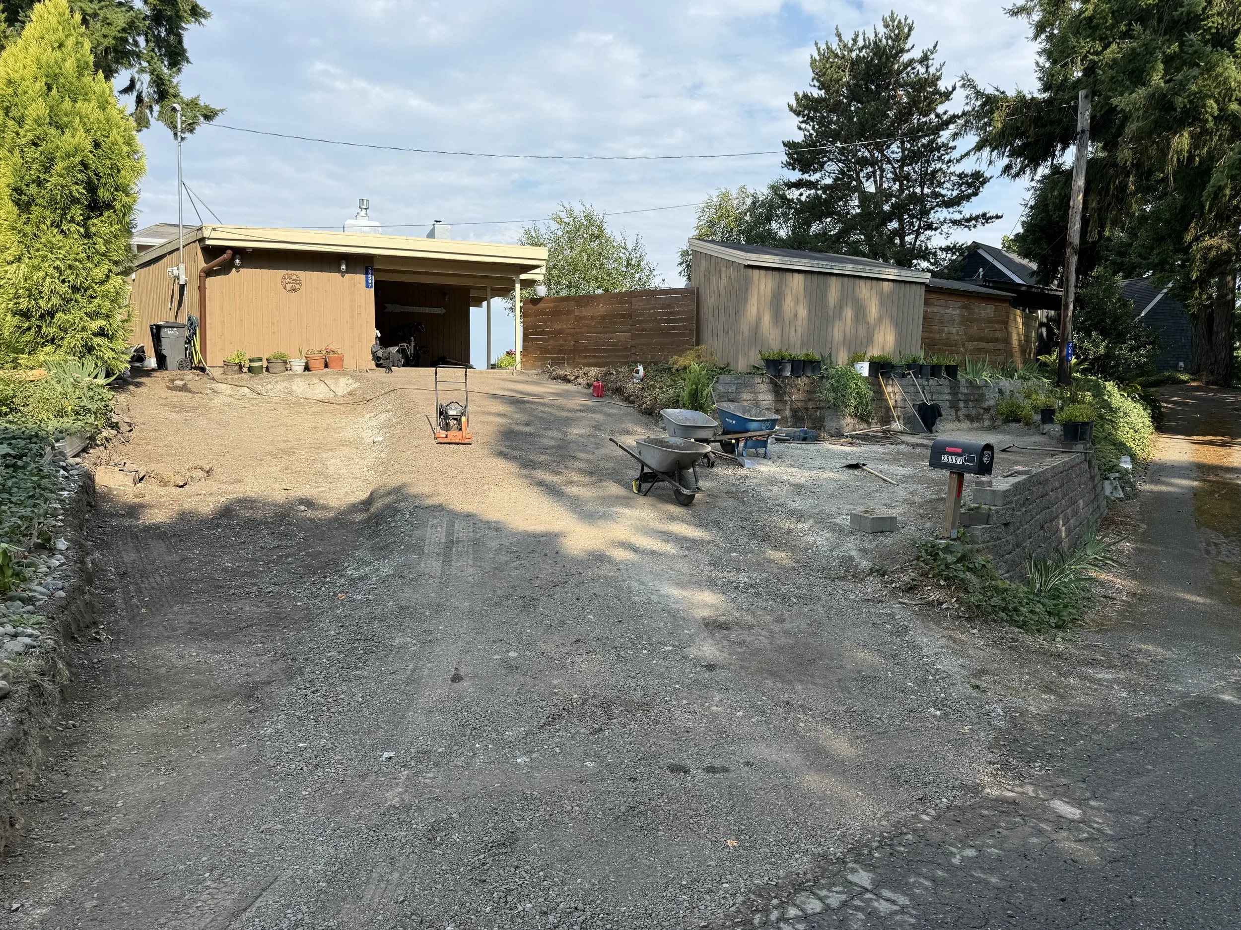 A driveway under construction with dirt and gravel, surrounded by trees and houses, construction tools, and materials scattered around.