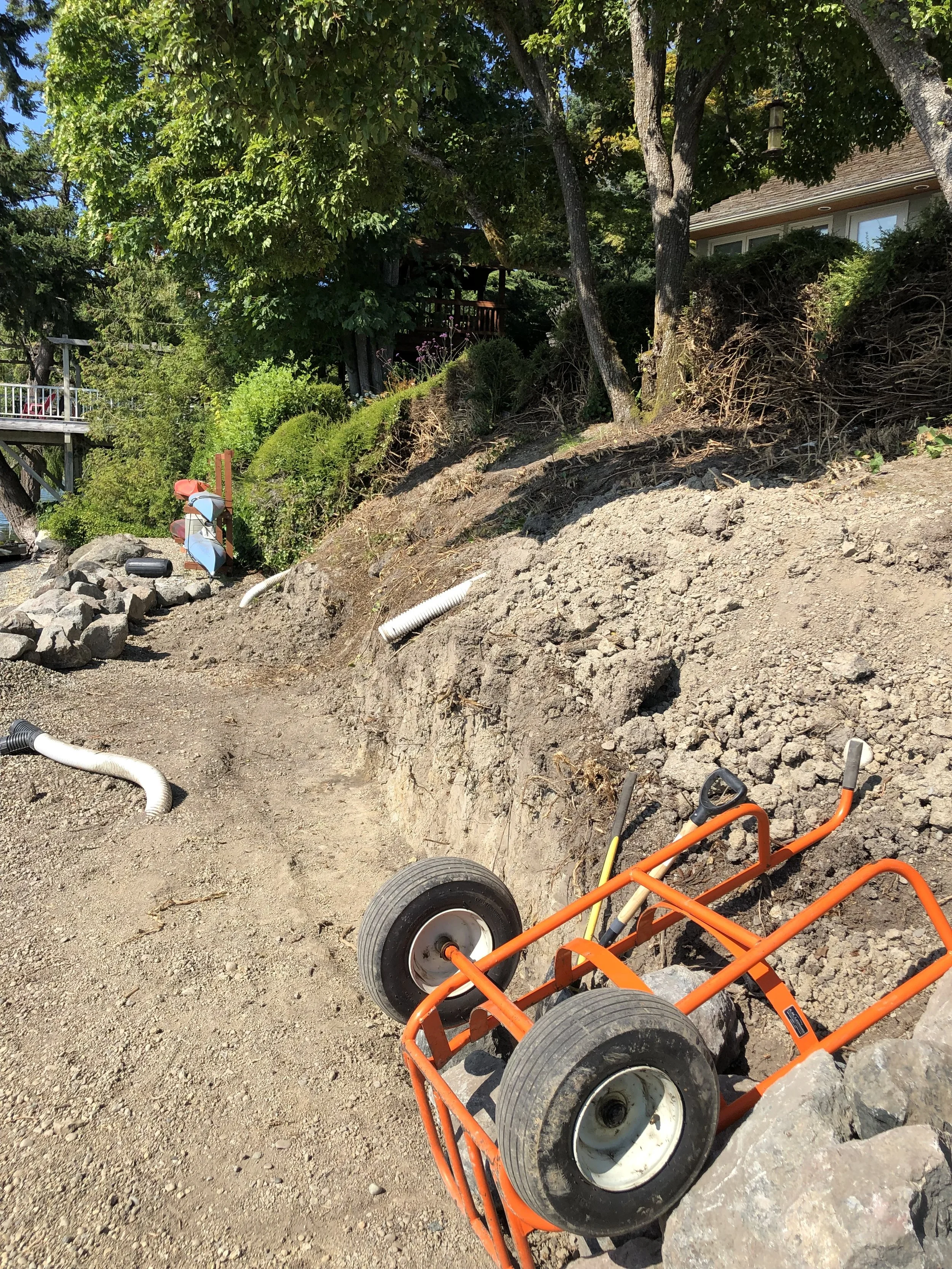 A garden or backyard area under construction with exposed dirt and rocks, a wheelbarrow, yellow and black work tools, white drainage pipes, lush green trees, shrubs, and bushes, with a house visible in the background.