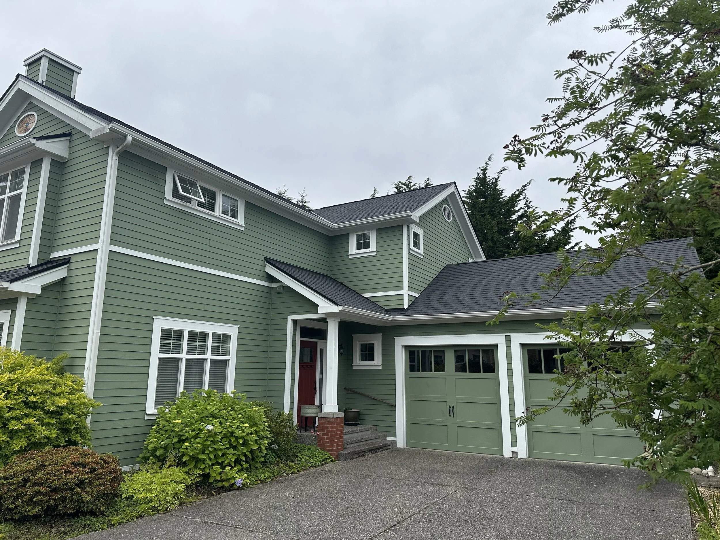 A two-story green house with white trim and a red front door, surrounded by greenery and trees, under a cloudy sky.