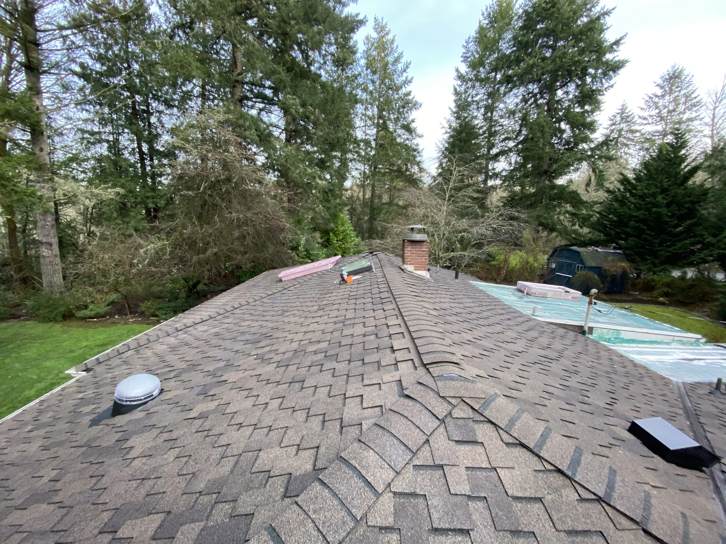 Residential roof with asphalt shingles, vents, and a chimney, overlooking trees and a neighboring house with a backyard area.