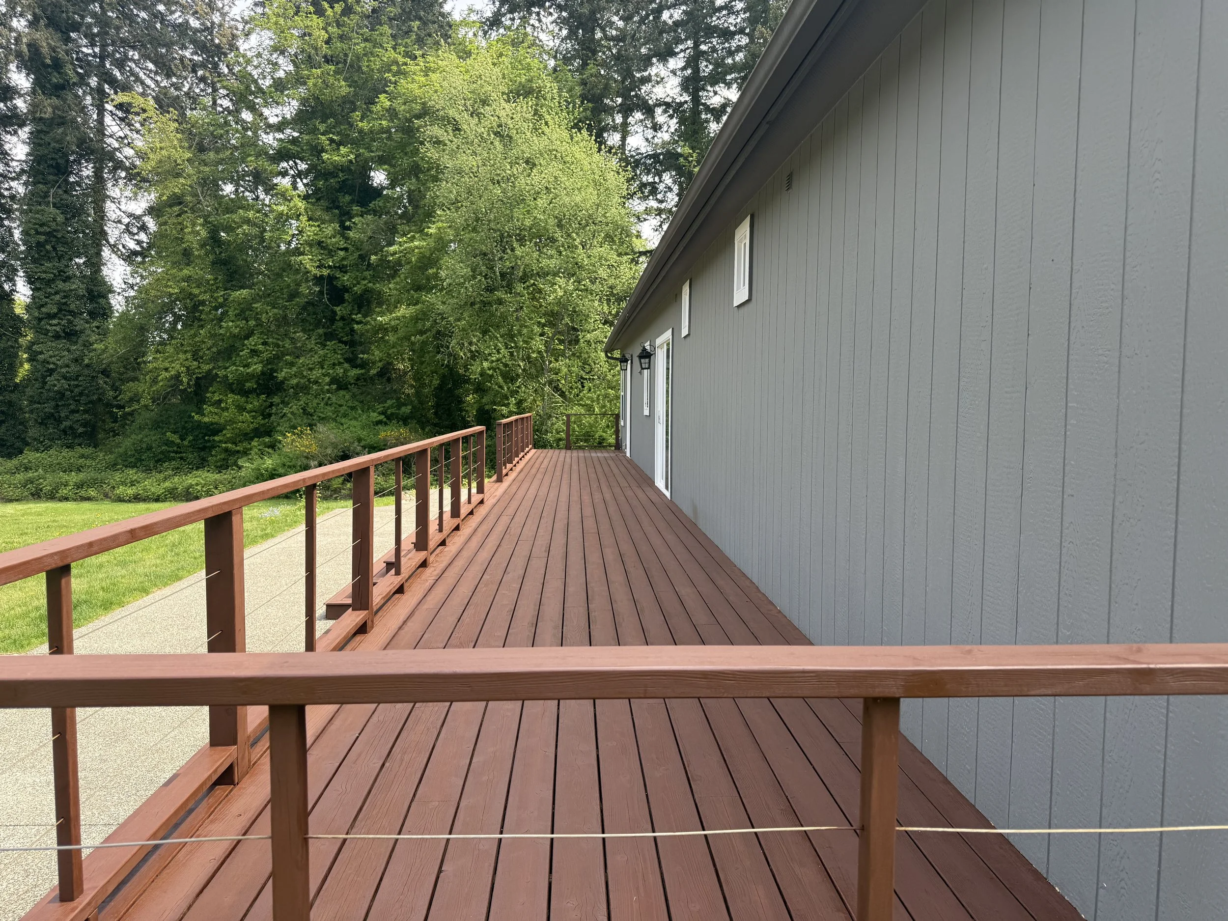 A wooden deck attached to a house with gray siding, overlooking a lush green yard and surrounded by tall trees.