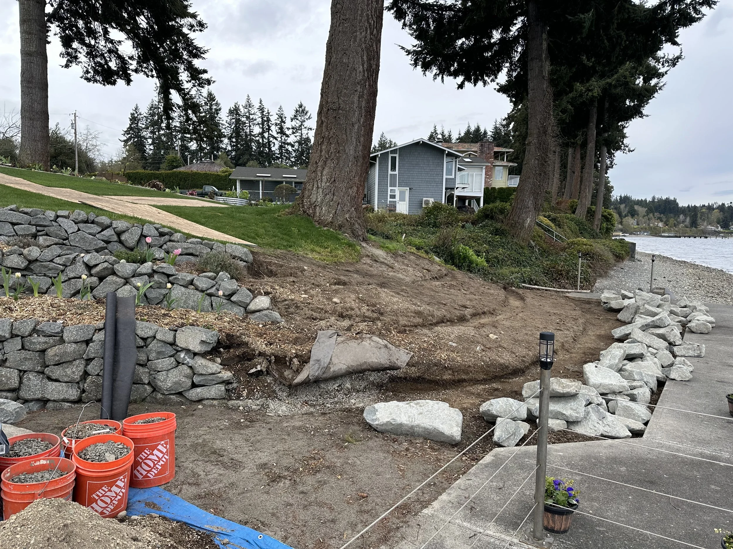 Outdoor landscaping or yard work in progress along a waterfront with trees and houses in the background, and construction materials like orange buckets, rocks, and a blue tarp in the foreground.