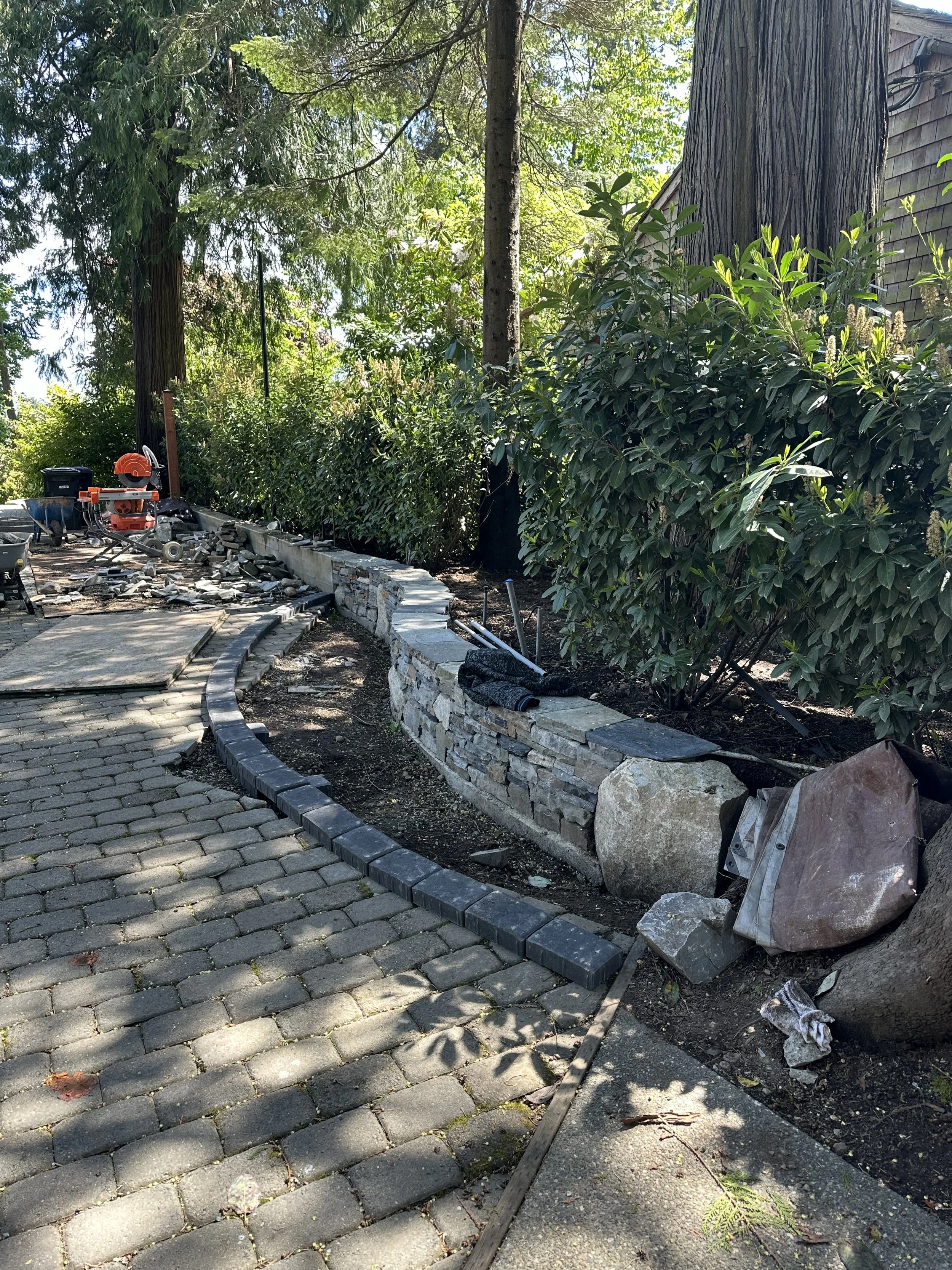 A landscaped garden area in process of being built or renovated. It shows a curved pathway made of cobblestone pavers, with a newly constructed low stone wall alongside it. Construction tools and materials, including a saw, are scattered near the wal