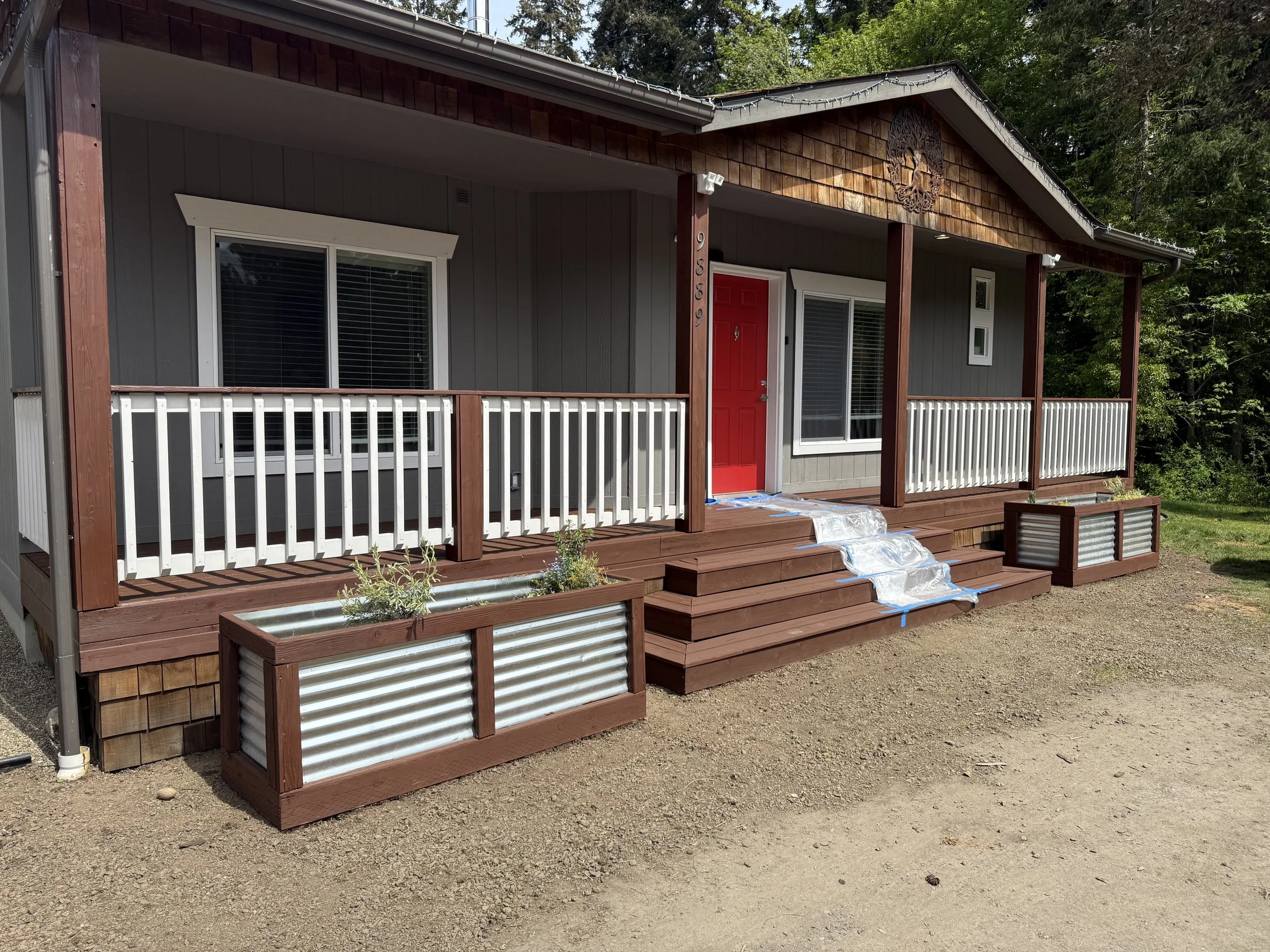 Front view of a small house with a reddish front door, gray siding, white railing on the porch, and decorative wooden and metal elements, surrounded by trees.
