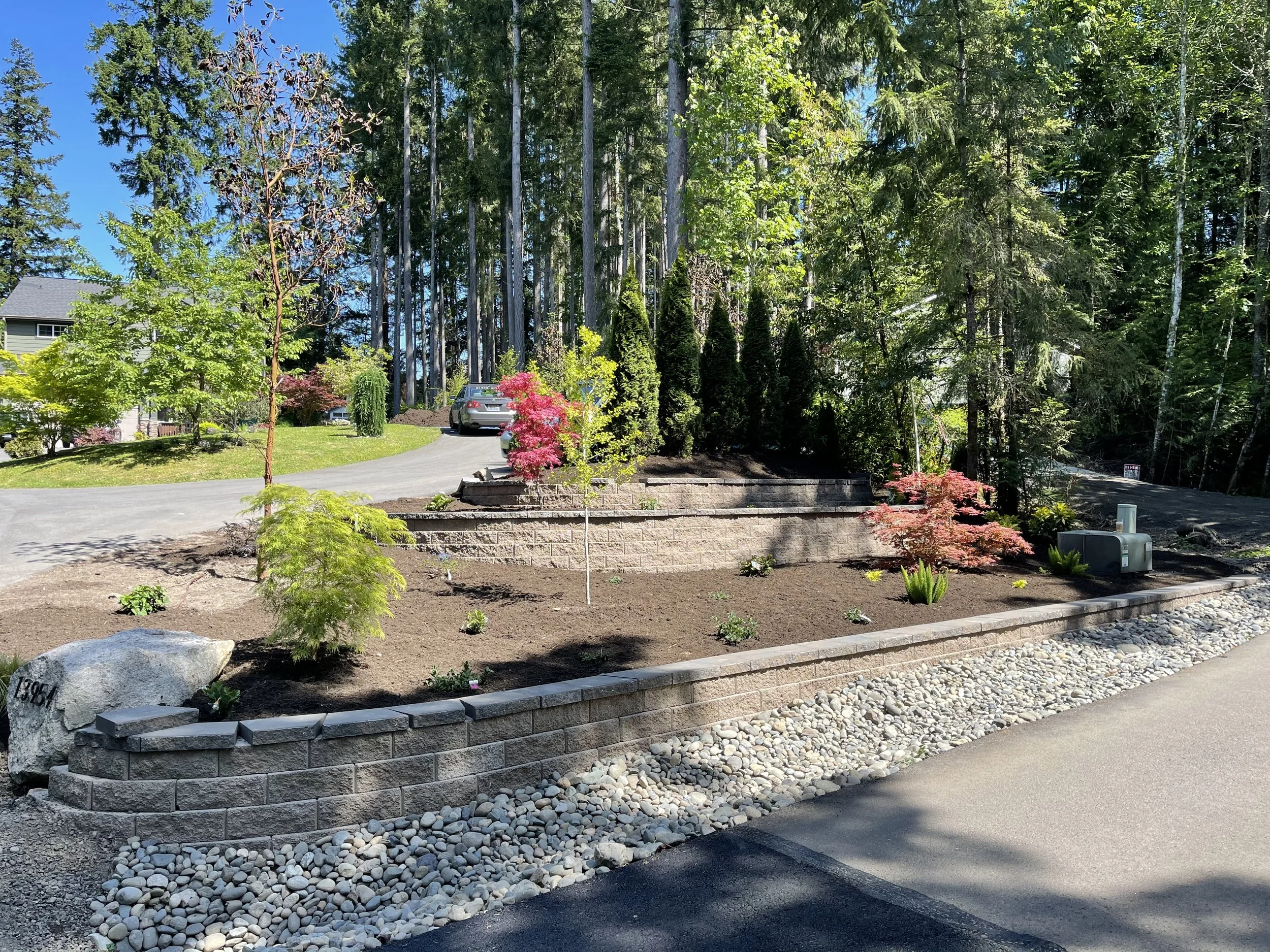 A landscaped yard with a raised garden bed bordered by gray bricks, filled with small plants and soil. There are decorative rocks and pebbles along the sidewalk, a variety of trees, bushes, and a house with a gray roof visible in the background, unde