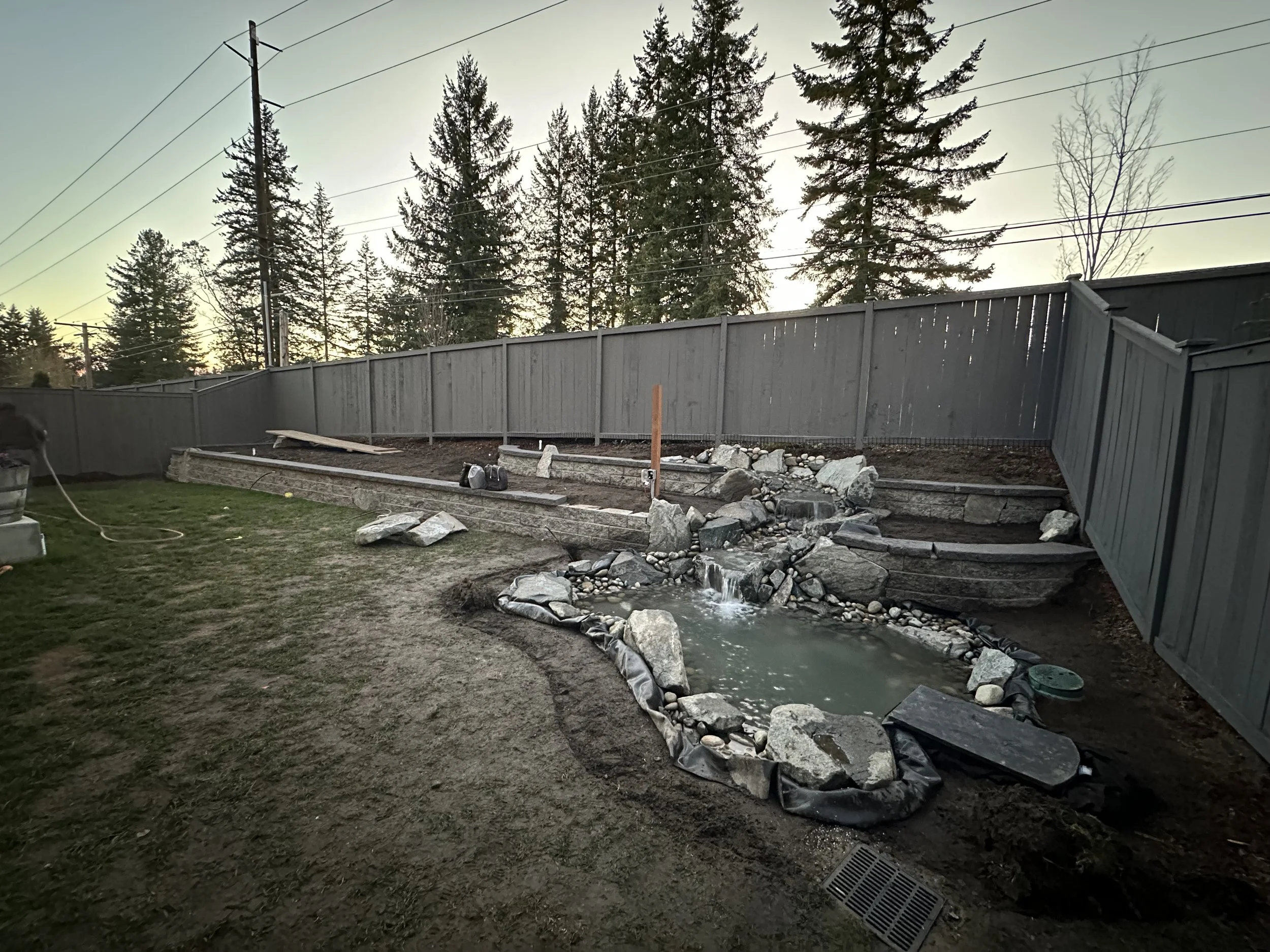 A backyard with a gray wooden fence under a partly cloudy sky, trees in the background, and a small stream of water flowing over rocks in the foreground, with some construction or landscaping debris present.