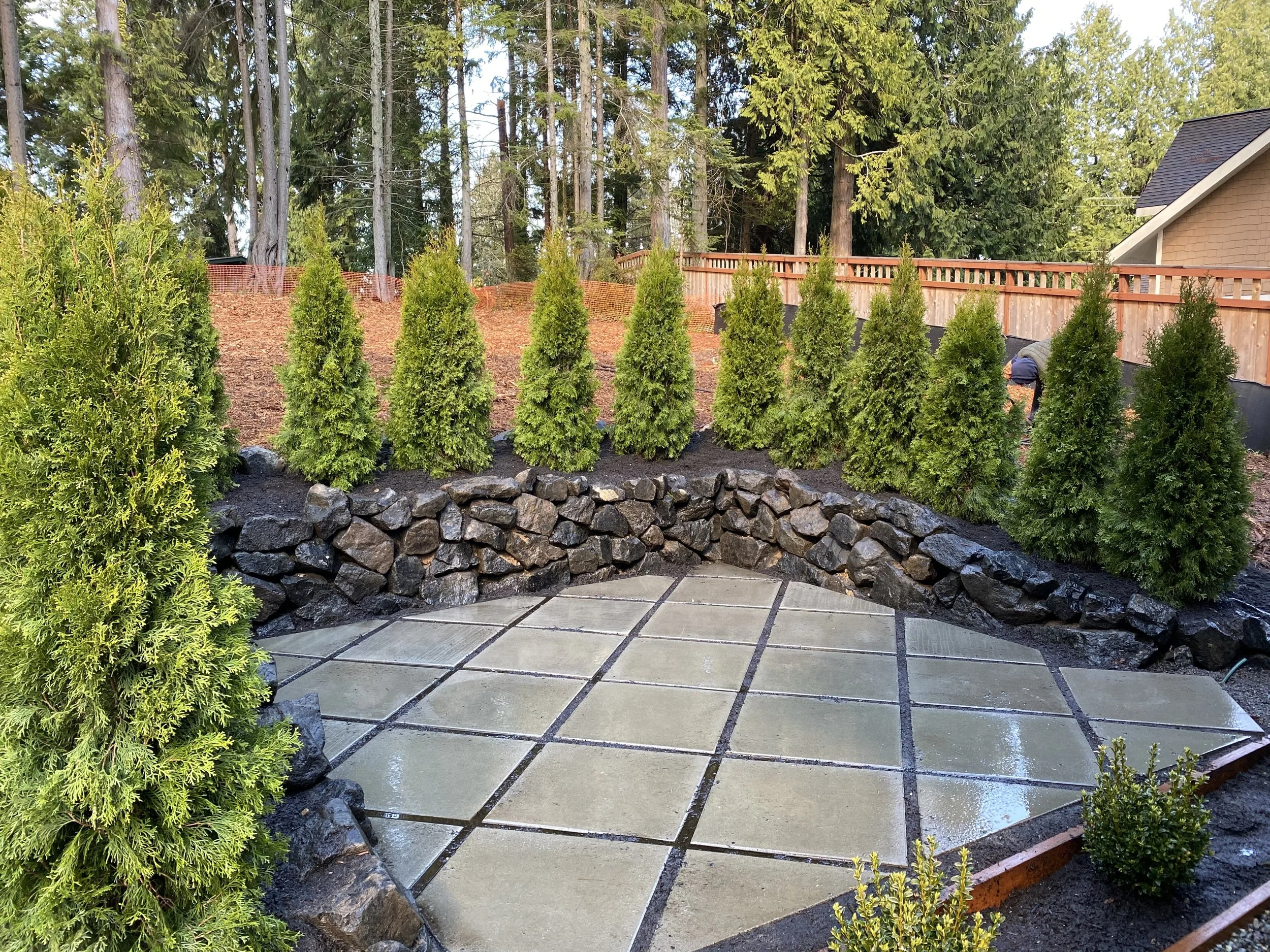Backyard with newly installed paved patio, stone edging, green bushes, and a wooden fence with tall trees in the background.