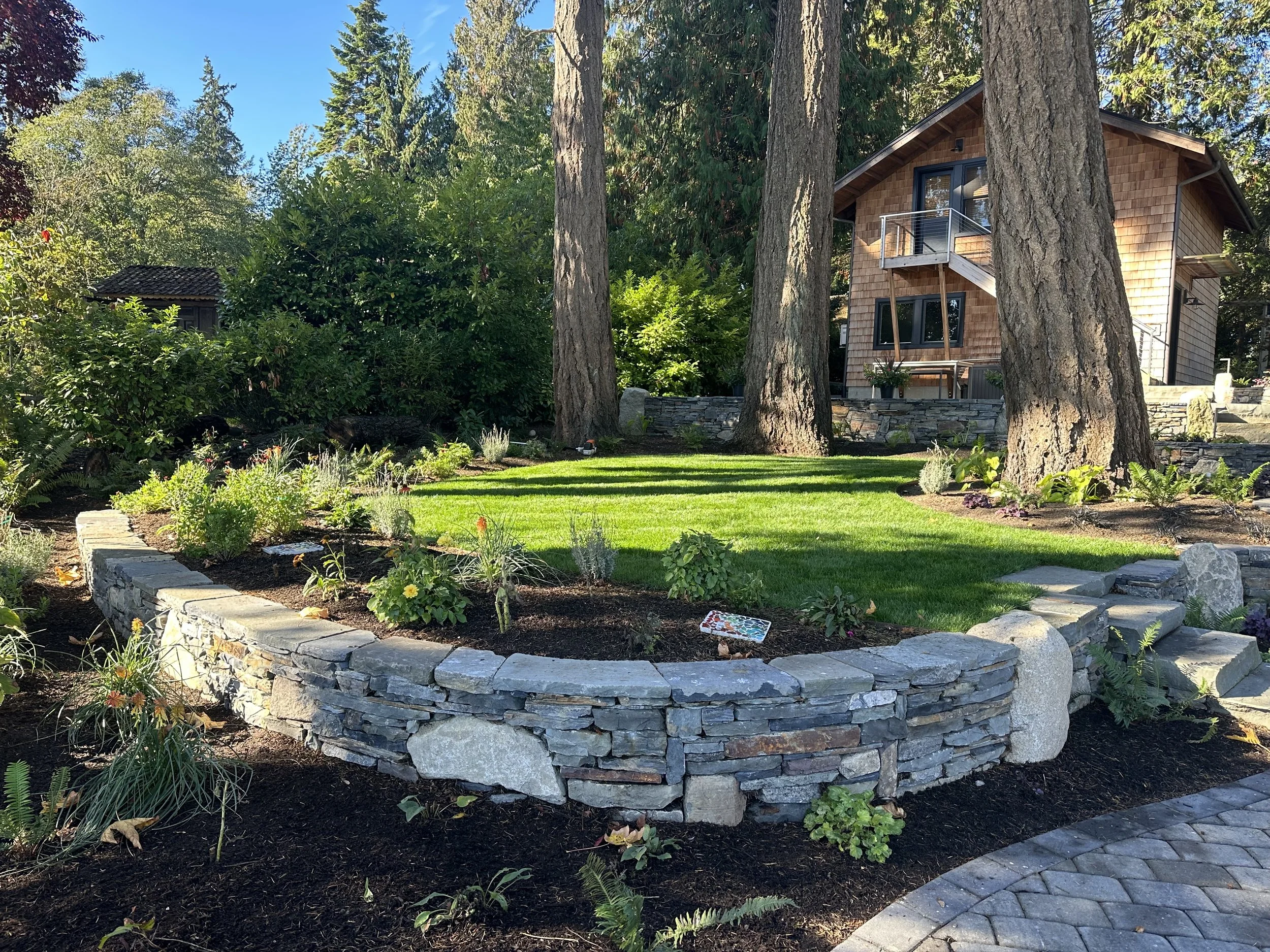 A landscaped backyard with a stone patio, lush green grass, flowering plants, and tall trees surrounding a modern house.