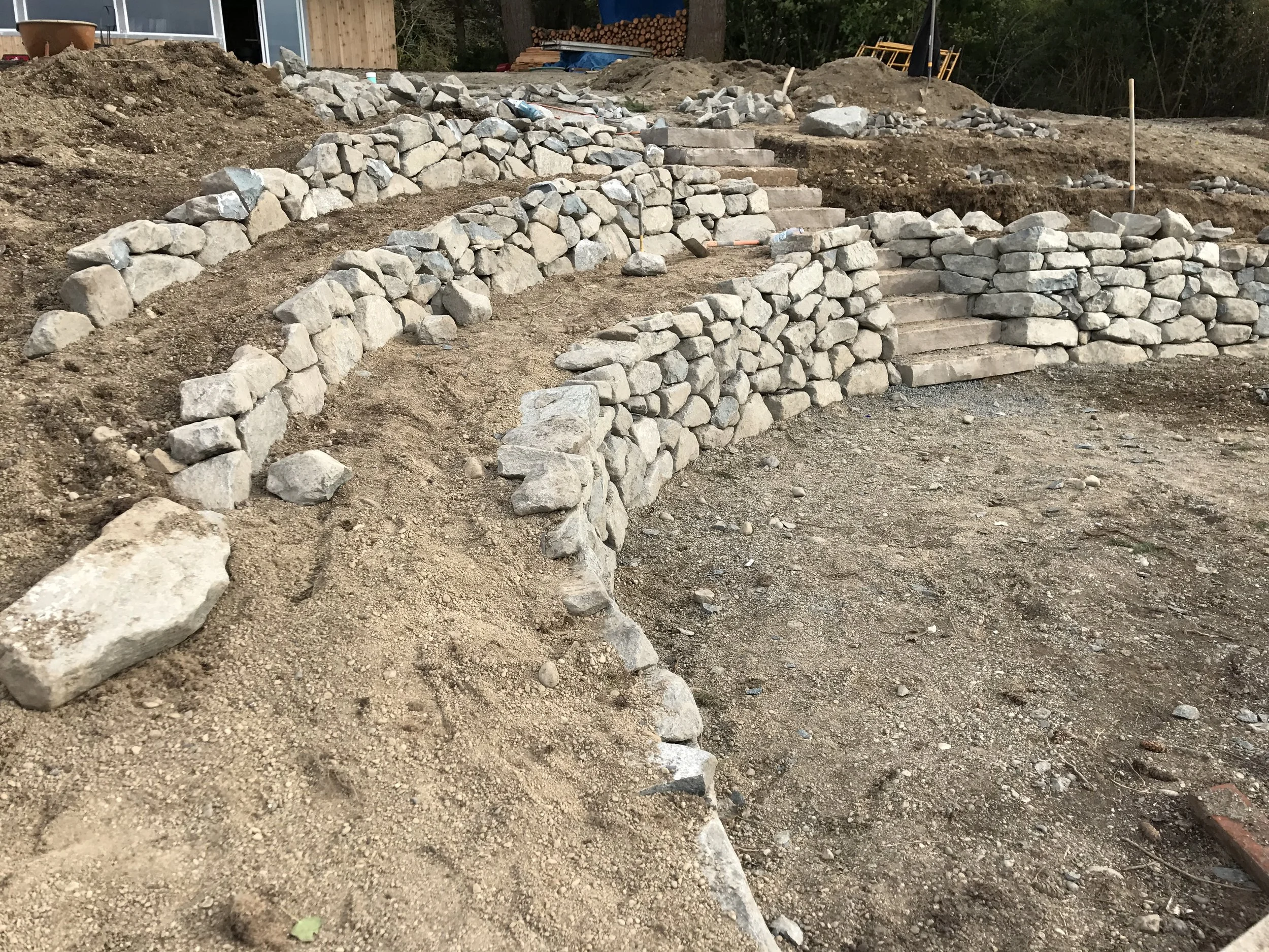 A backyard construction site with stone steps being built and rocks stacked along the sides, dirt and tools scattered around.