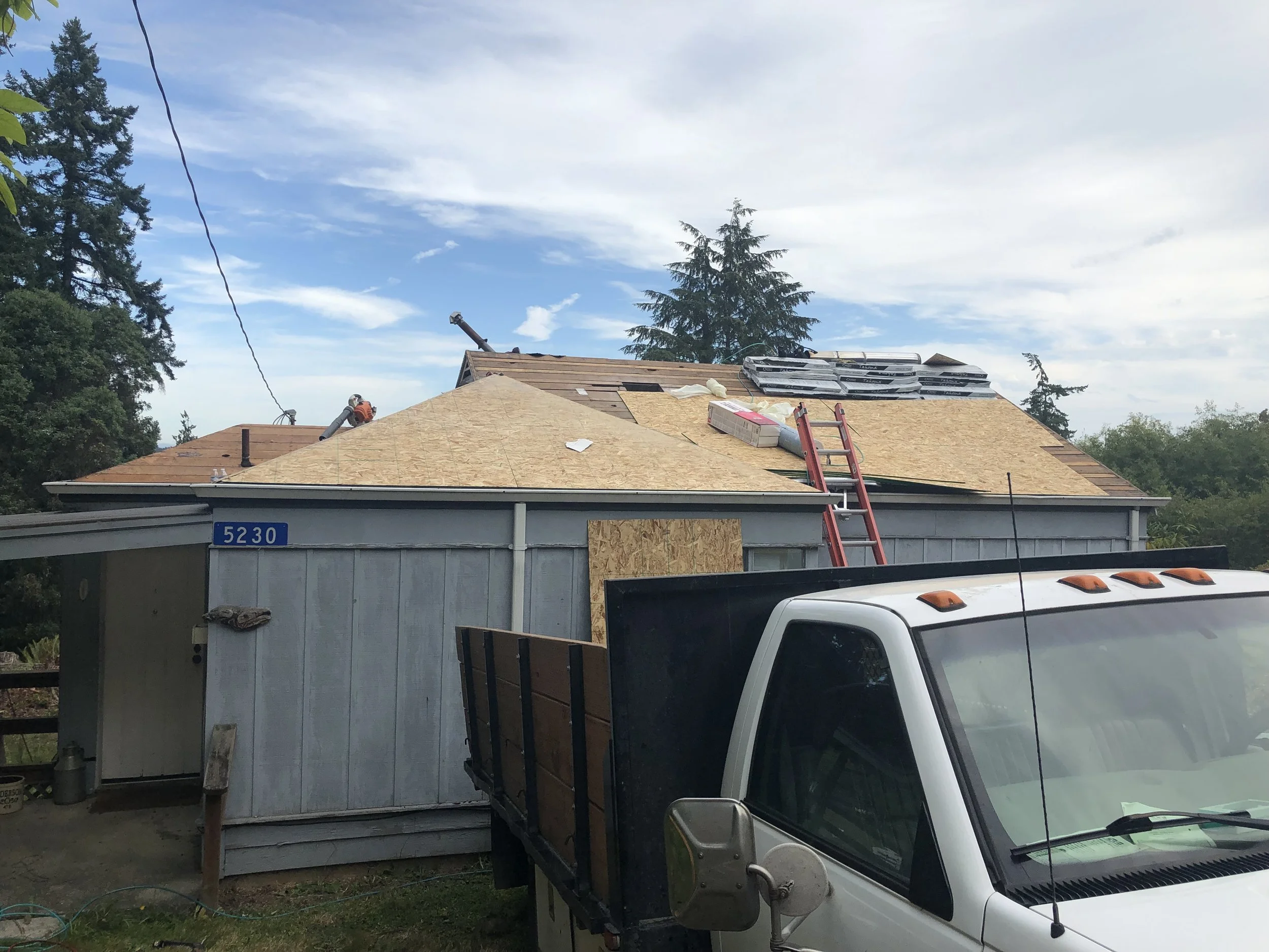 A house under construction with a partially shingled roof, a wooden panel on the side, a ladder leaning against it, and construction tools and materials visible. Tall trees and a cloudy sky in the background.
