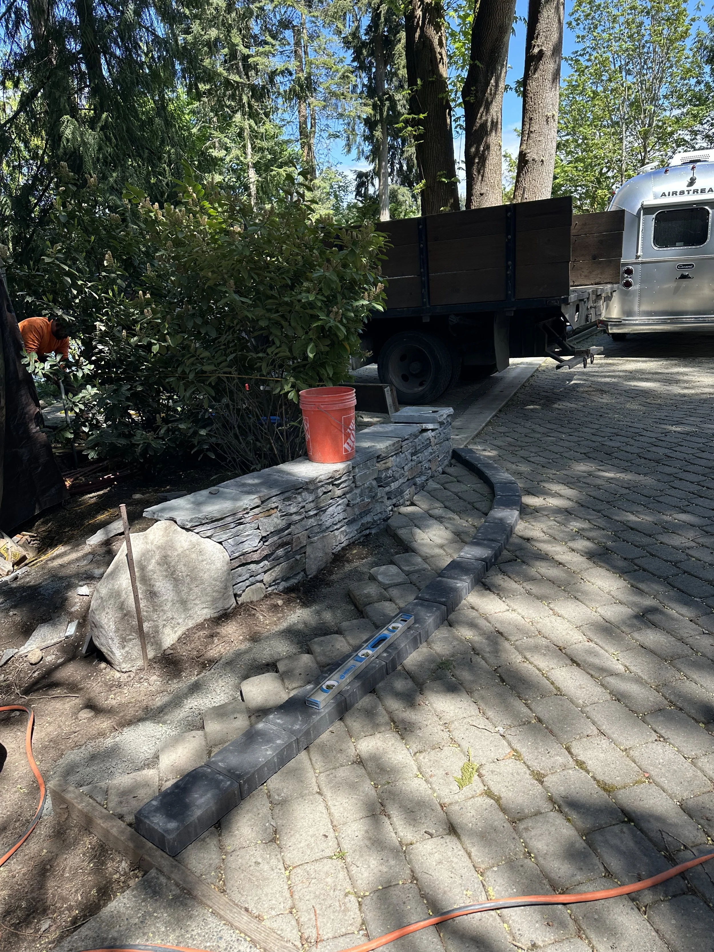 A construction worker is working on landscaping or outdoor construction, with a truck and trailer nearby, and an assortment of stones and pavers being laid on a sidewalk or patio area.