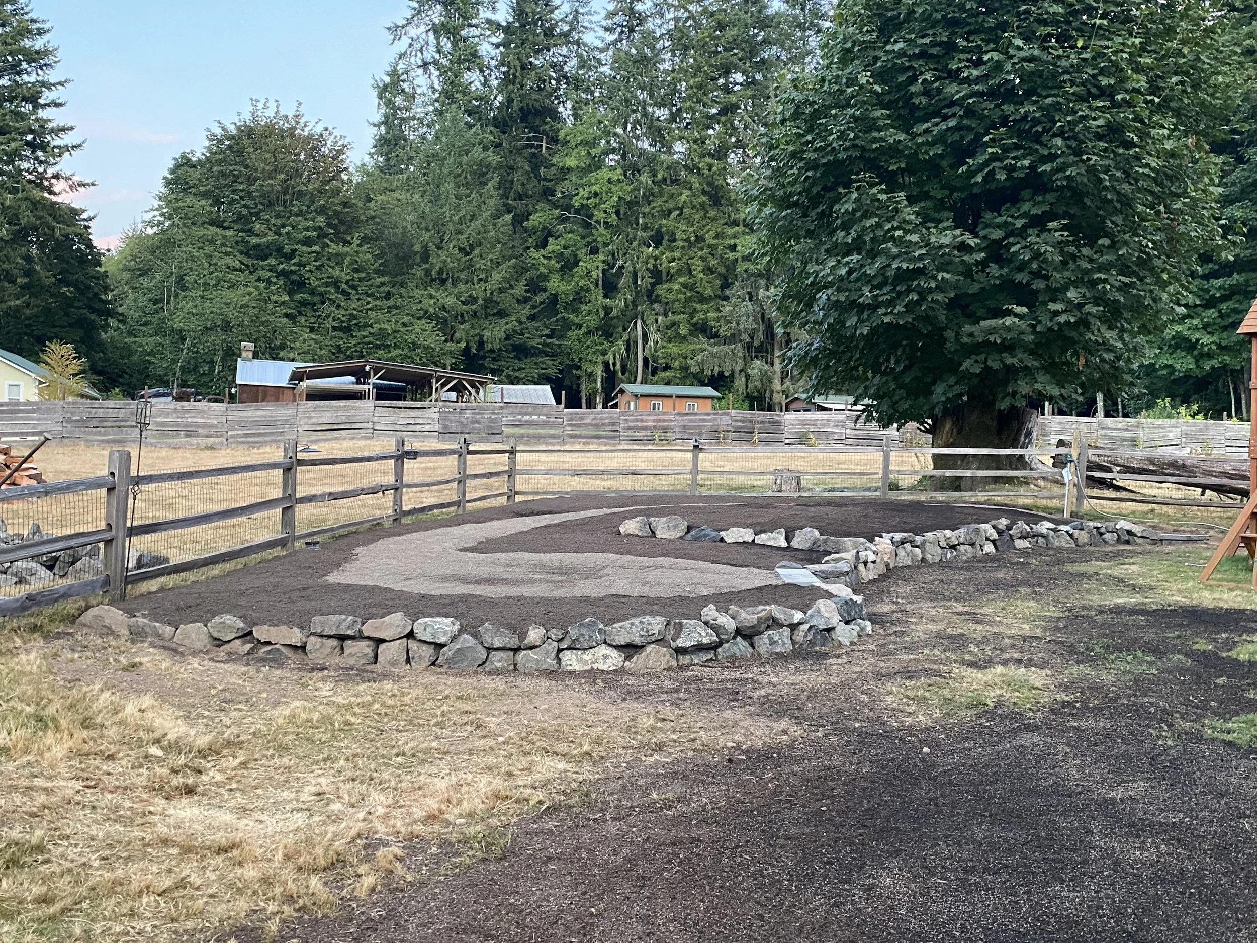 A fenced yard with a circular stone border, dry grass, large trees in the background, and small buildings or sheds with metal roofs.