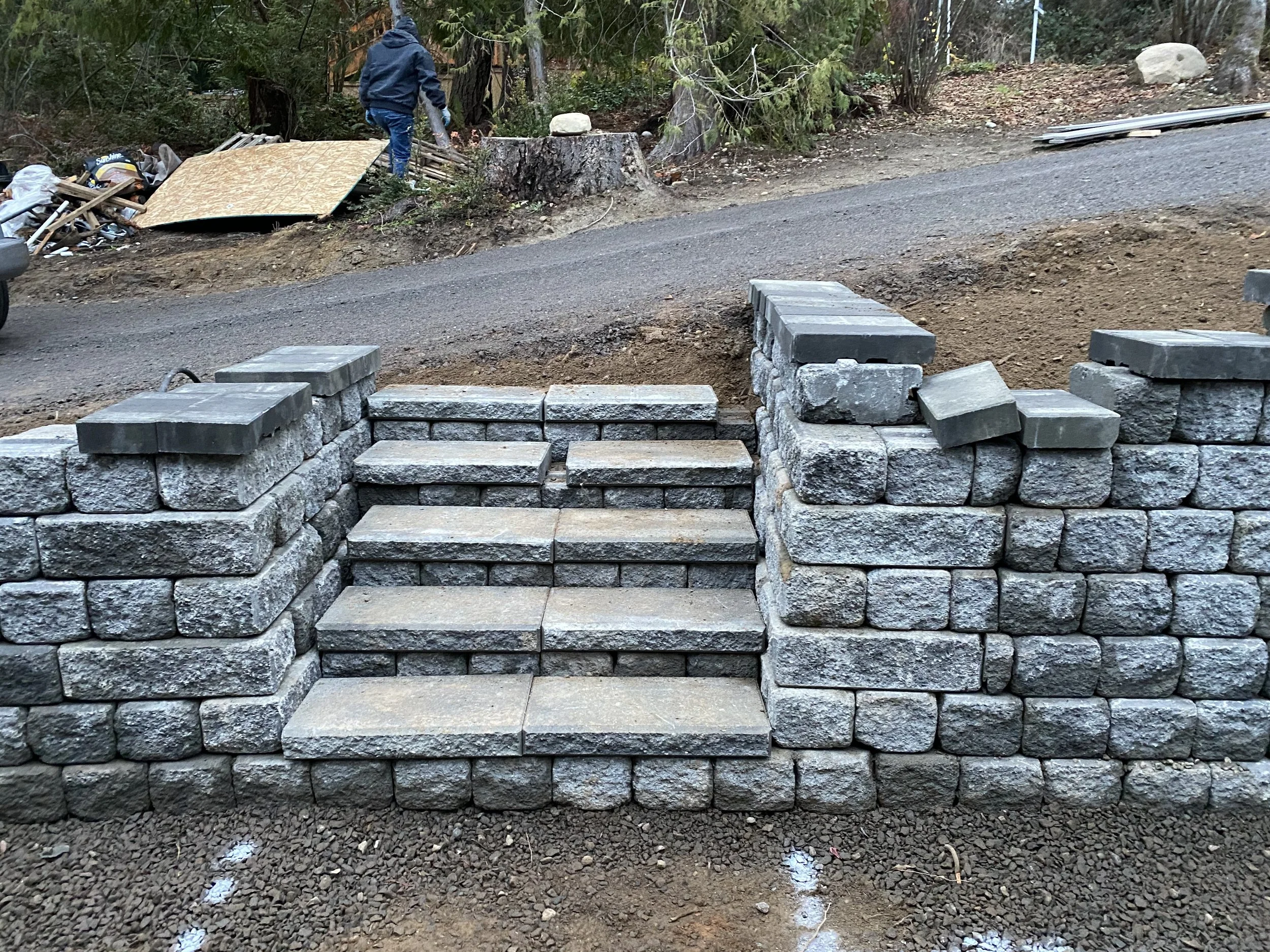 Construction worker building a stone retaining wall in a natural outdoor setting with trees and dirt.