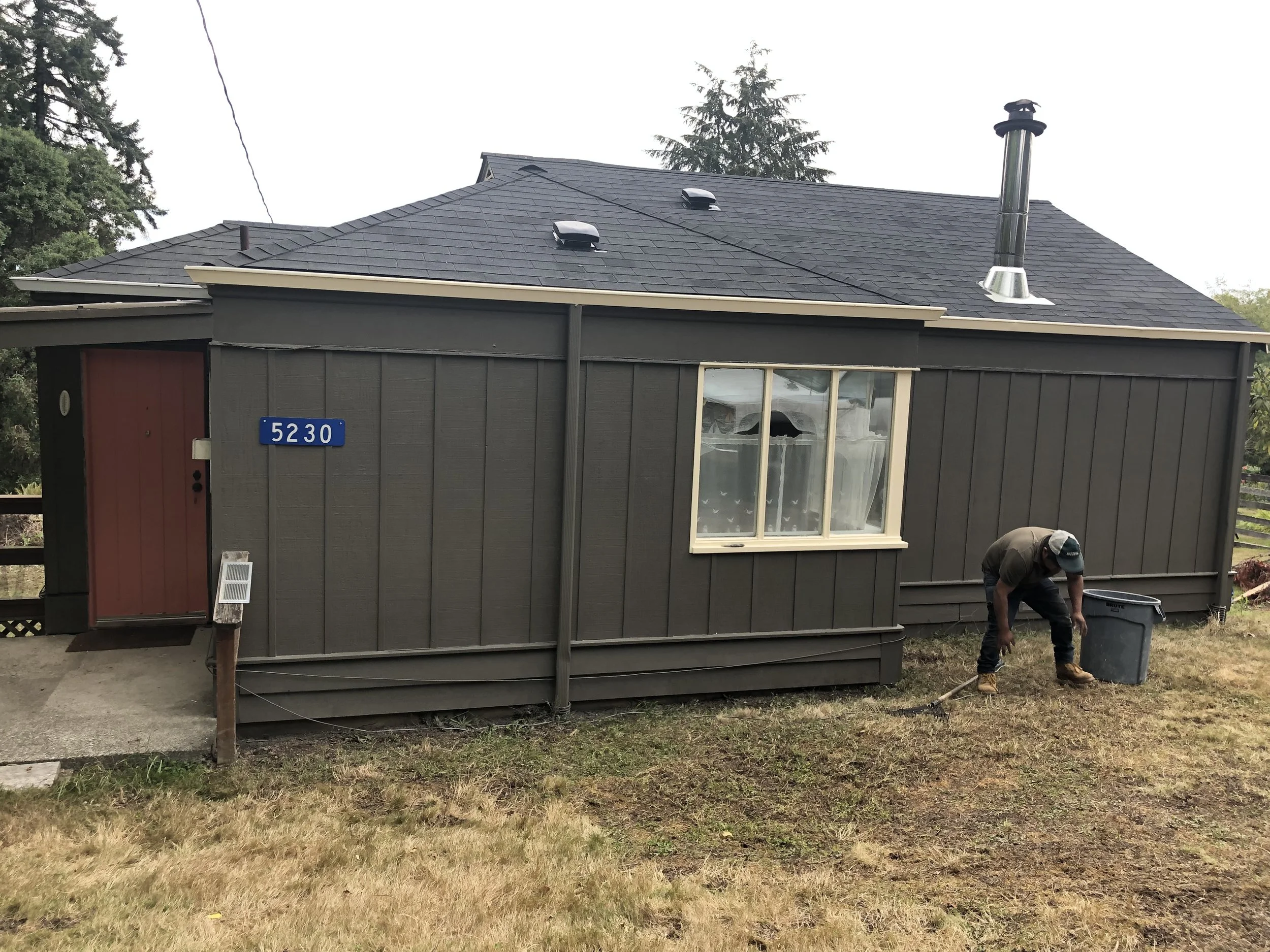 The exterior of a small house with dark gray siding, a red door, and a white-framed window. The house has a gray shingle roof with two vents and a chimney. There is a house number sign reading '5230' on the front. A person is crouched on the grass in