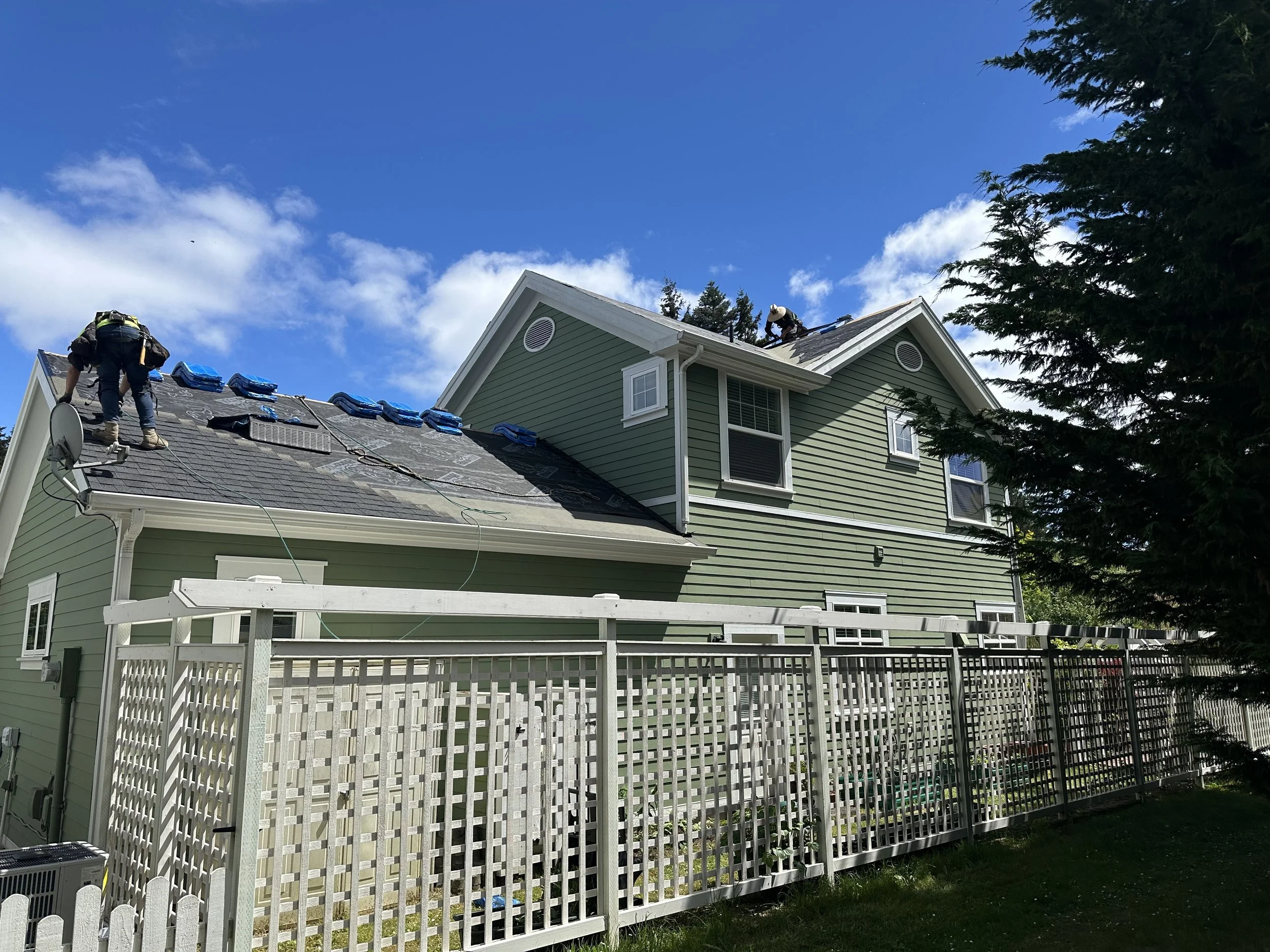 Two workers on a roof replacing shingles, one standing near the gutter and the other on the rooftop; the house is green with white trim and a fenced backyard, with trees and a partly cloudy blue sky in the background.