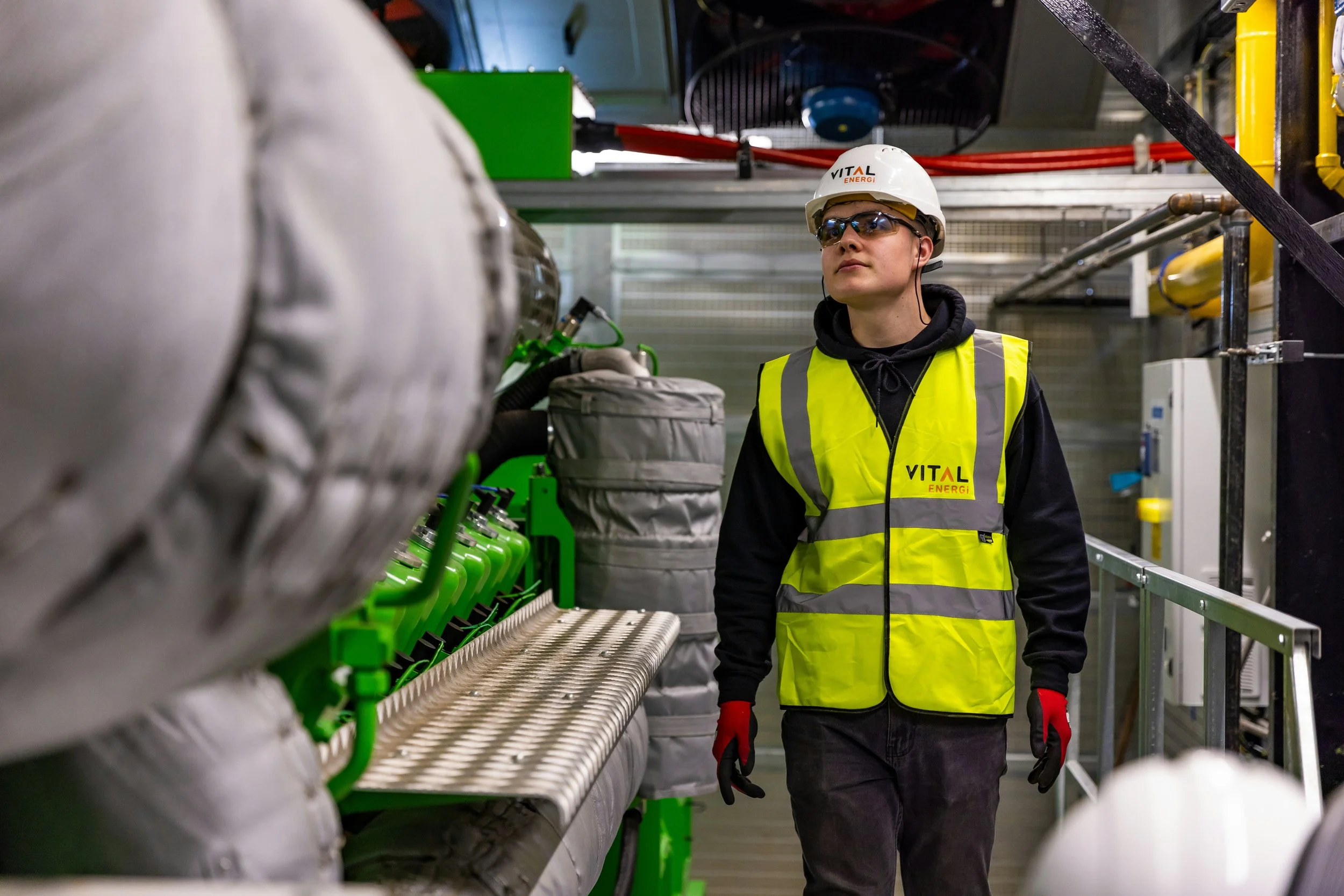 A person wearing safety glasses, a white hard hat with 'VITAL ENERGI' logo, a yellow high-visibility vest with 'VITAL ENERGI' logo, black hoodie, black pants, and red gloves working on industrial equipment in a factory or energy facility.