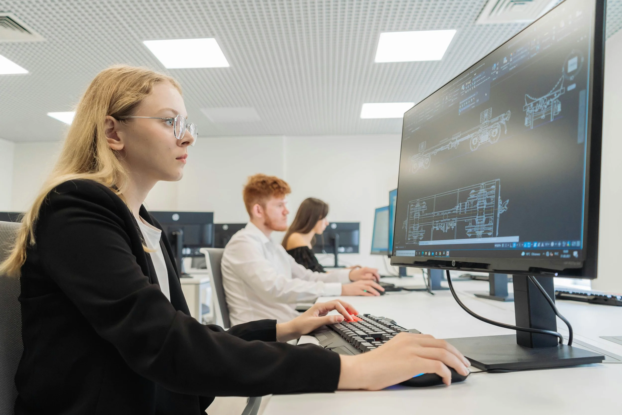 A woman with blonde hair and glasses working on a computer with technical blueprints on the screen in an office environment with other colleagues in the background.