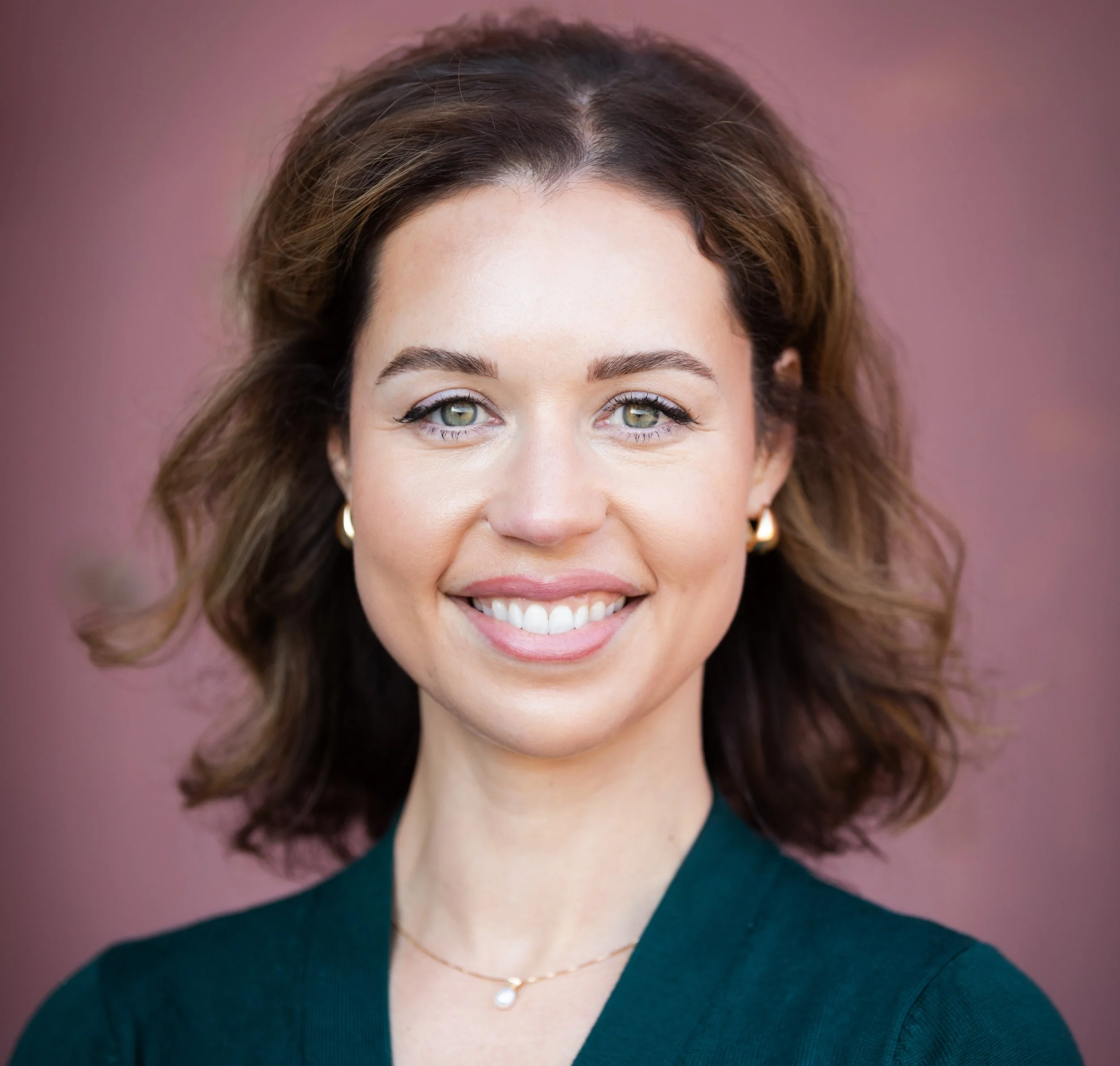 A picture of Erin Guthrie. Curly brown hair, wearing a dark teal top, gold hoop earrings, and a gold necklace with a pearl pendant, against a pink background.