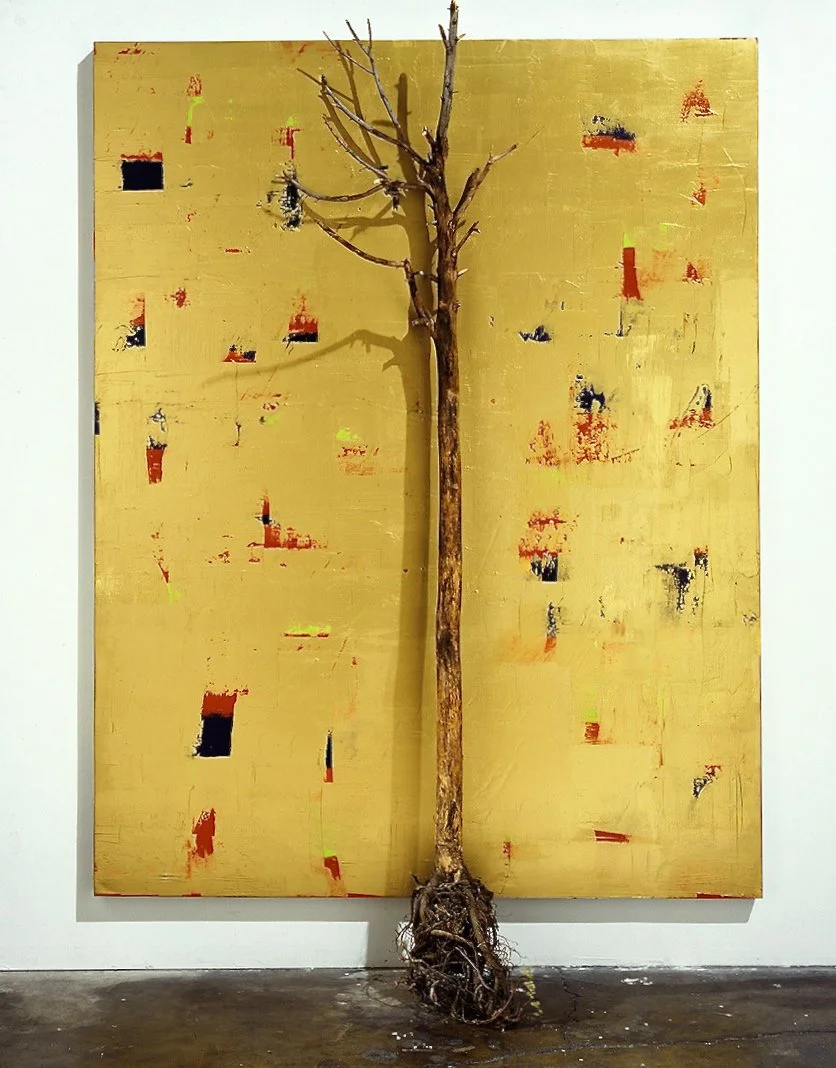 A stick-shaped tree with roots at the bottom is mounted in front of an Abstract yellow and red painting on a wall.