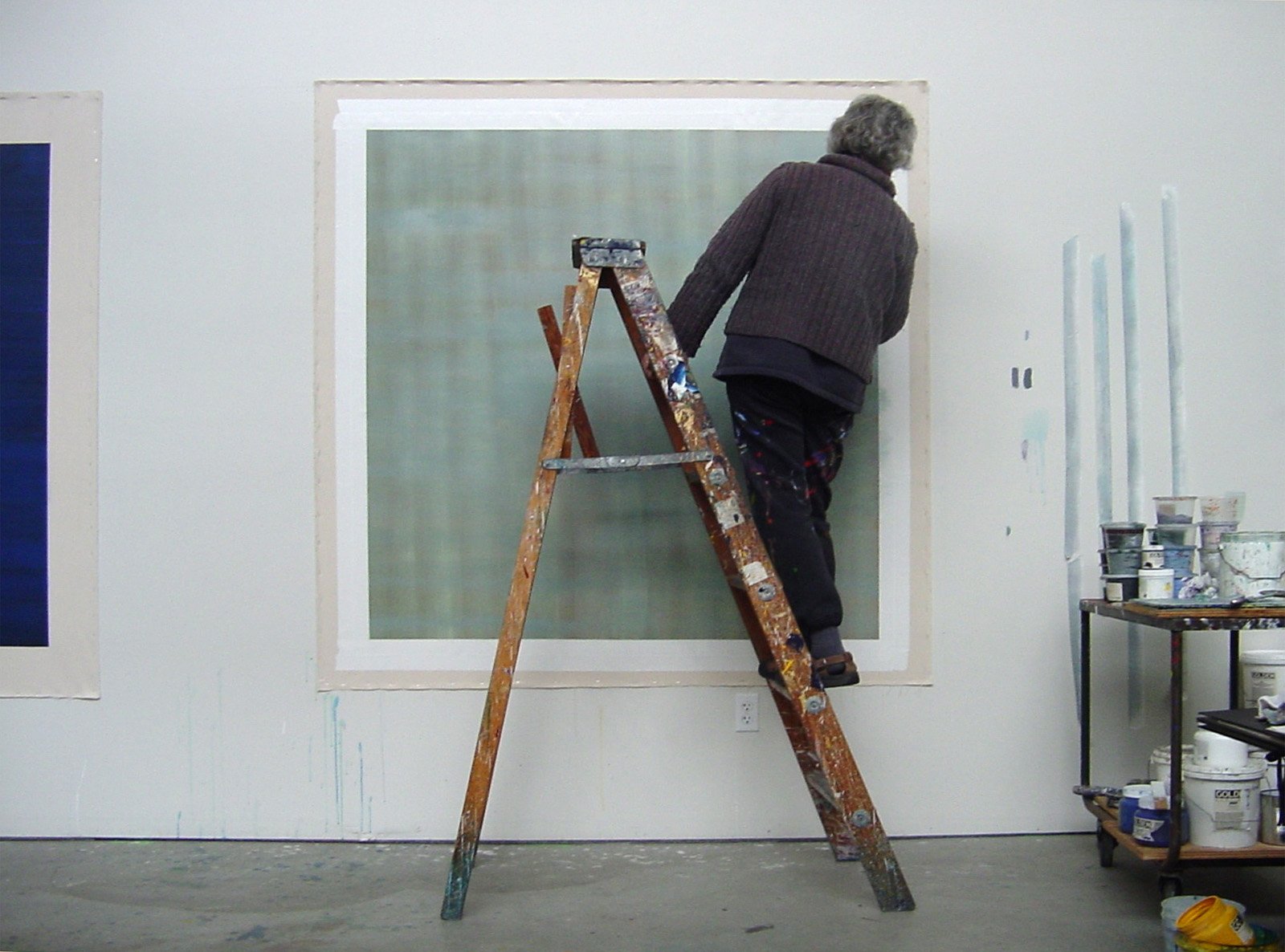 An elderly person is standing on a ladder, painting a wall with a large roller brush. There are paint supplies on a cart nearby.