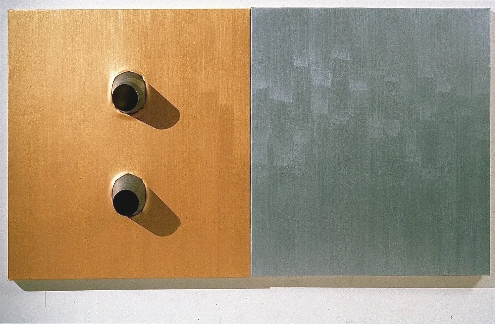 Two cylindrical objects are mounted on a flat surface, with shadows cast to the right. The surface has a two-tone color, with the left side being a warm beige and the right side a muted gray.