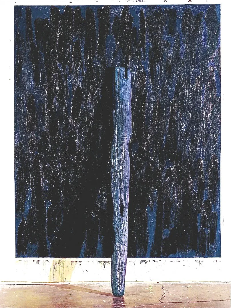A vertical, textured wooden sculpture stands in front of a large black and blue abstract painting on a white wall.