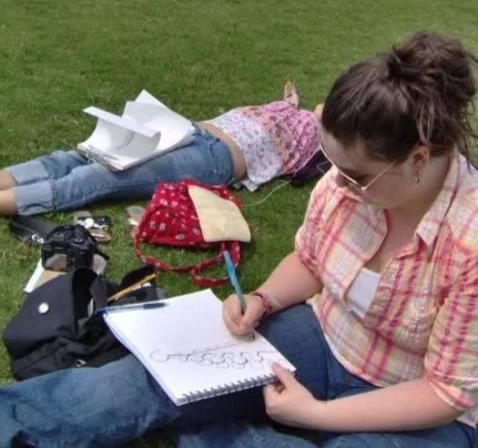 Woman drawing on a sketchpad while lying on grass, with a child lying face down in the background.