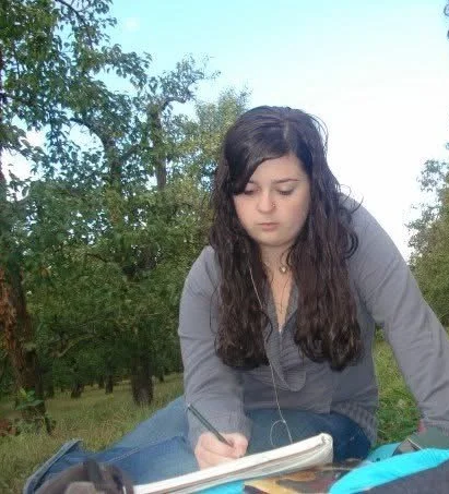 Young woman with long dark hair sitting outdoors, writing in a notebook with trees and blue sky in the background.