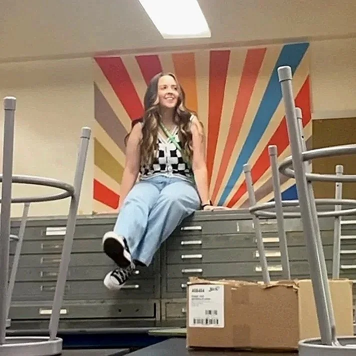 A girl sitting on a stack of chairs or tables in a room with a colorful striped mural on the wall behind her.