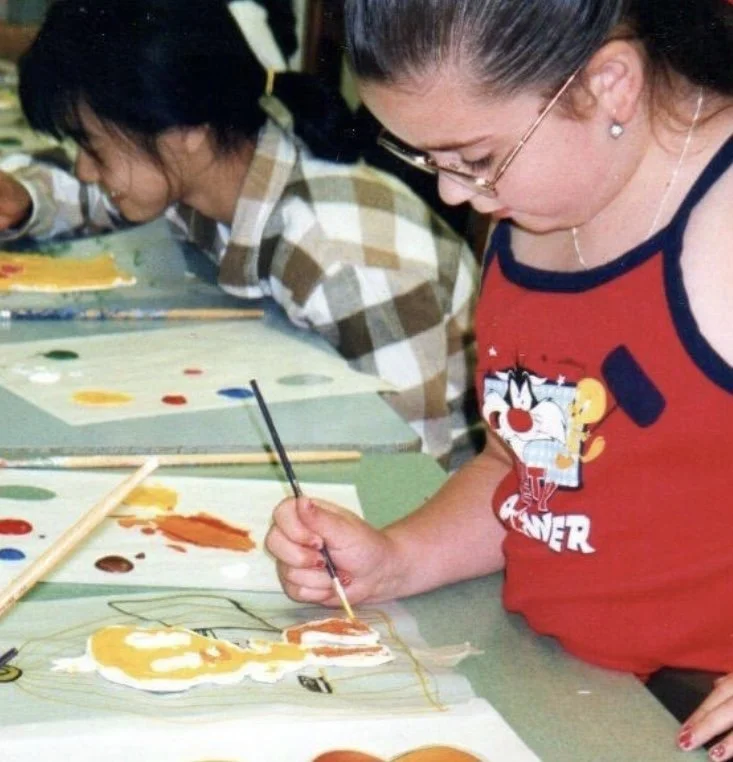 Two women painting on paper at a table, with various paint colors and brushes; one woman wearing glasses and a red tank top with cartoon character printing.