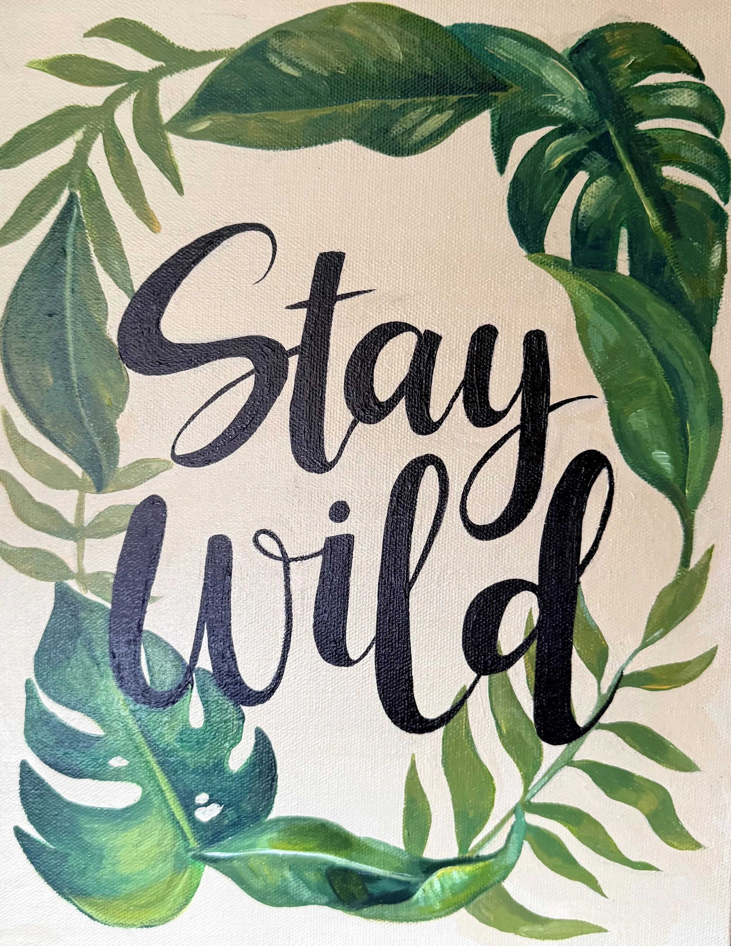 Close-up of a wall art with green tropical leaves surrounding black cursive text.